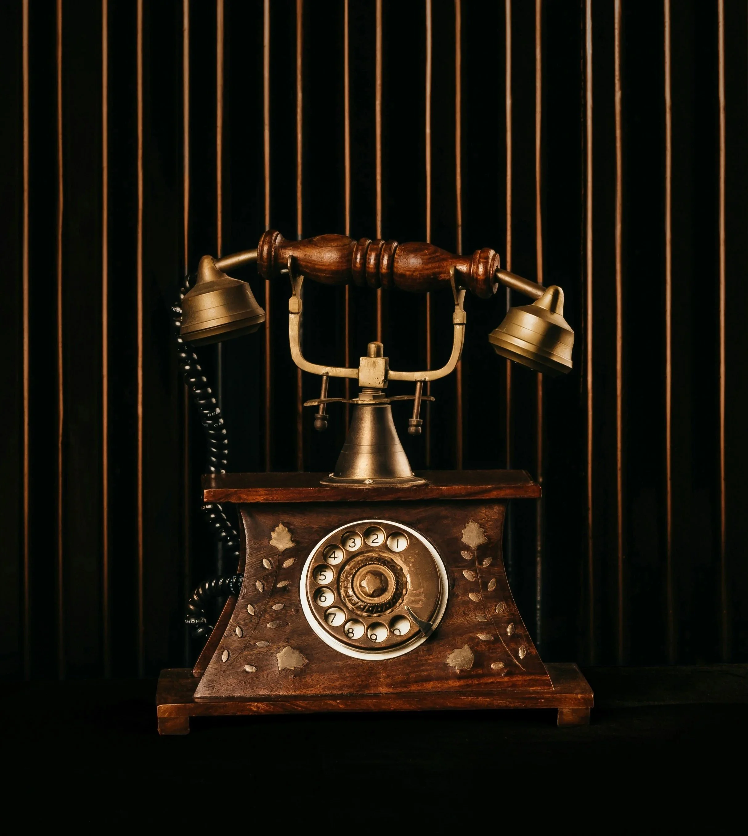 Vintage rotary dial telephone with a wooden base and brass accents, placed on a dark surface with a wooden backdrop and vertical orange-brown bars.