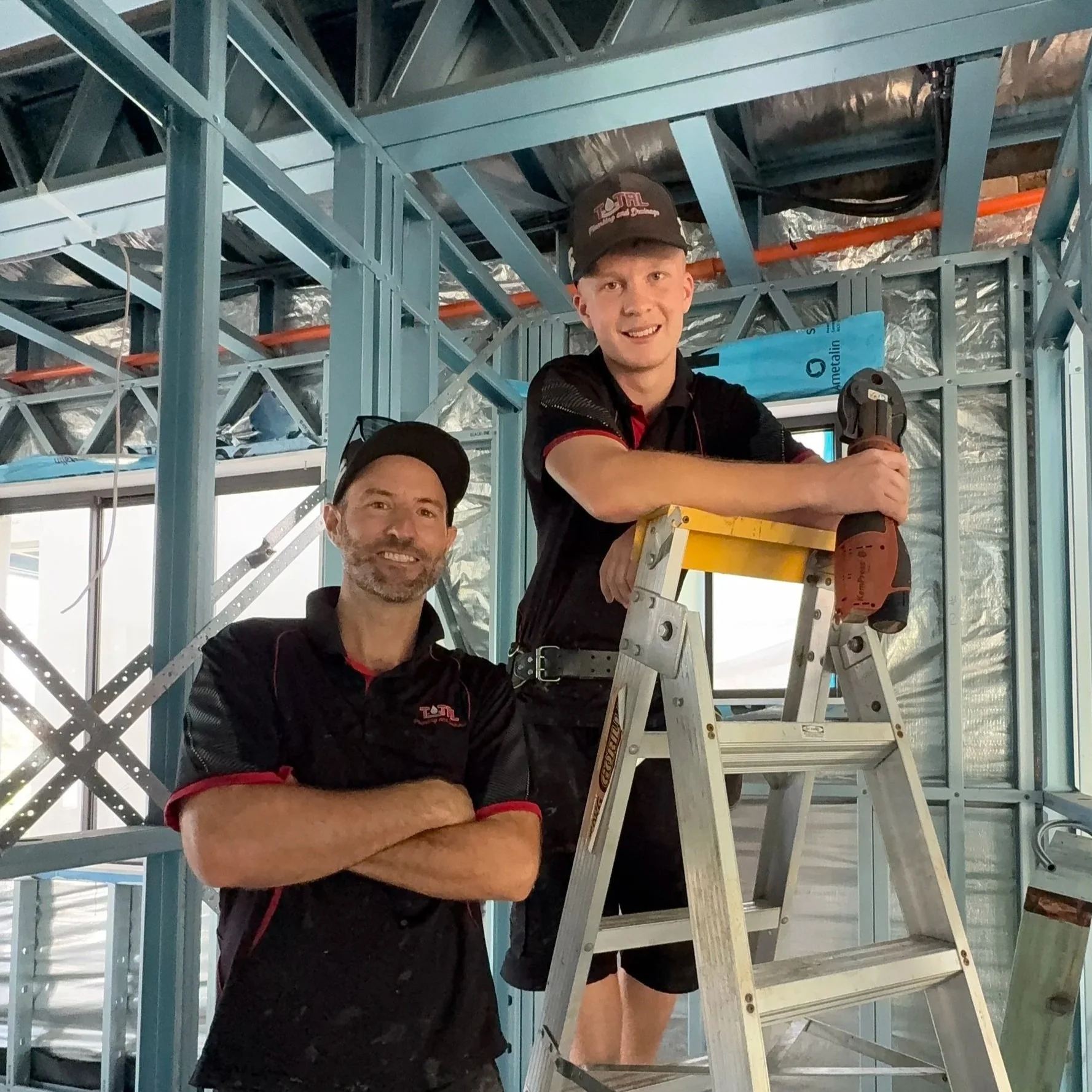 Two men in black uniforms working on a construction site with metal framing, one standing on a ladder holding a power drill, and the other standing next to him with arms crossed.