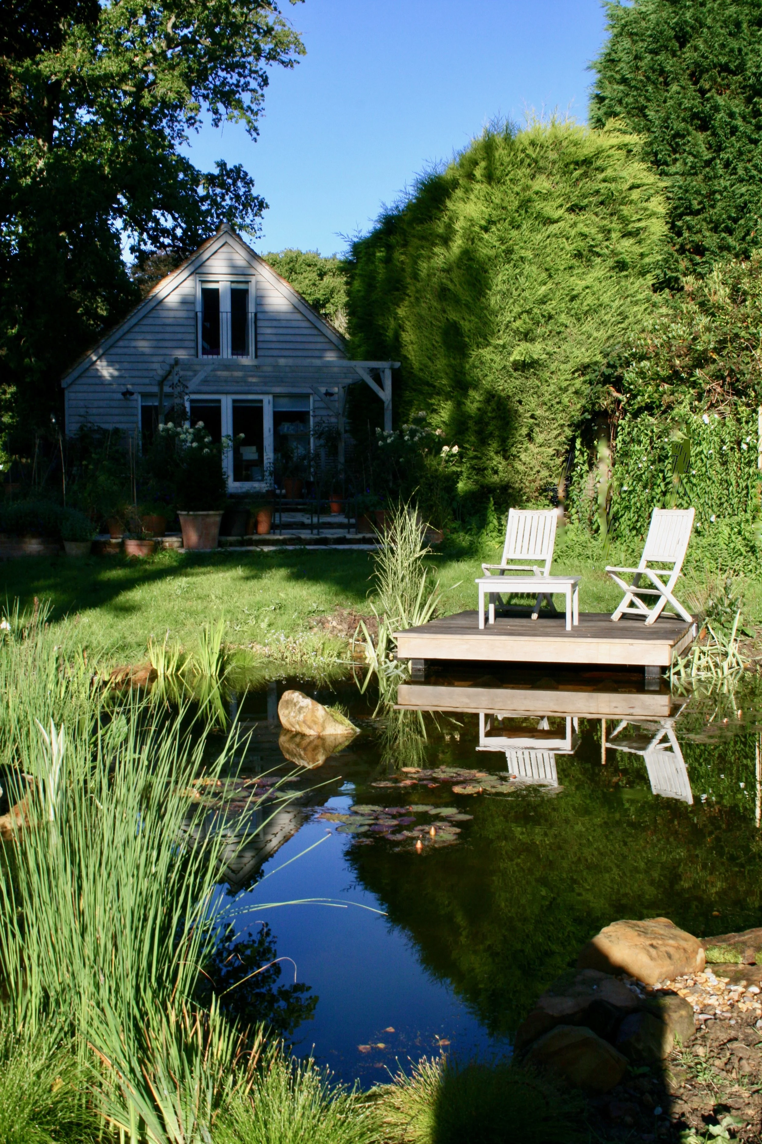 A garden with a pond, with a cantilevered  wooden deck, and a house with gray siding surrounded by green trees and bushes.