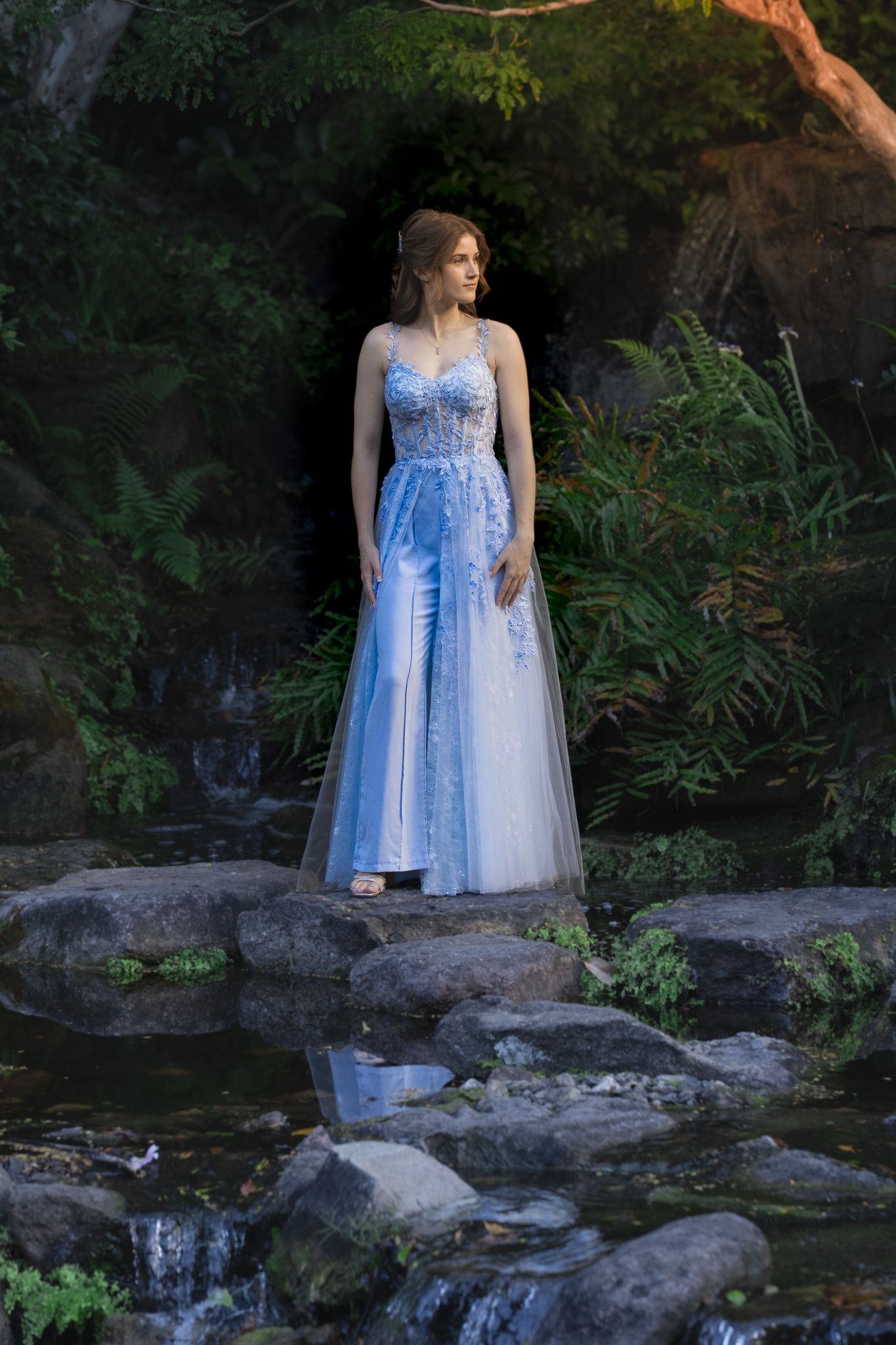 A woman in a detailed blue and white gown stands on rocks near a small waterfall in a lush, green forest setting.