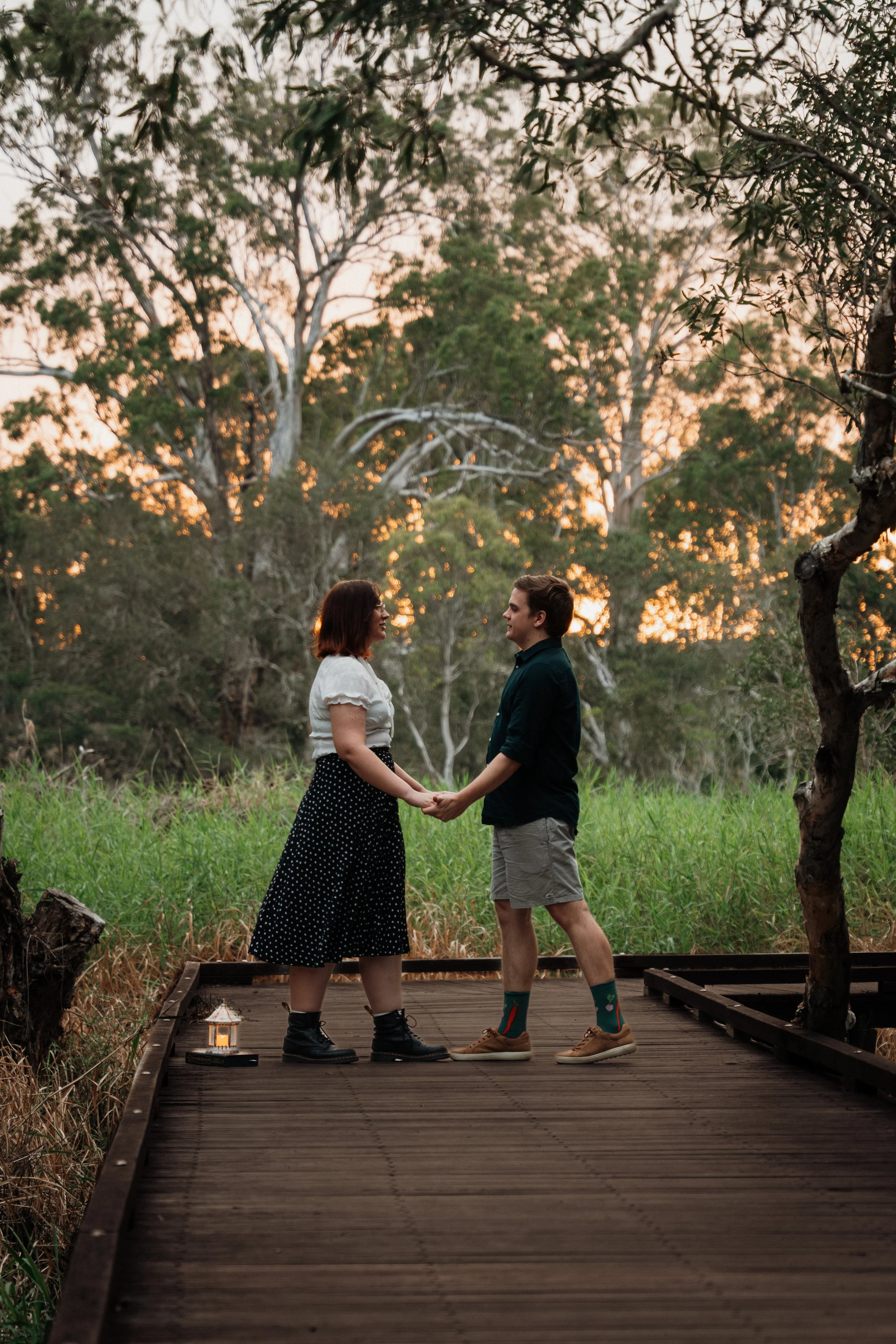 A couple stands on a wooden bridge, holding hands and facing each other, outdoors at sunset with trees and greenery in the background.