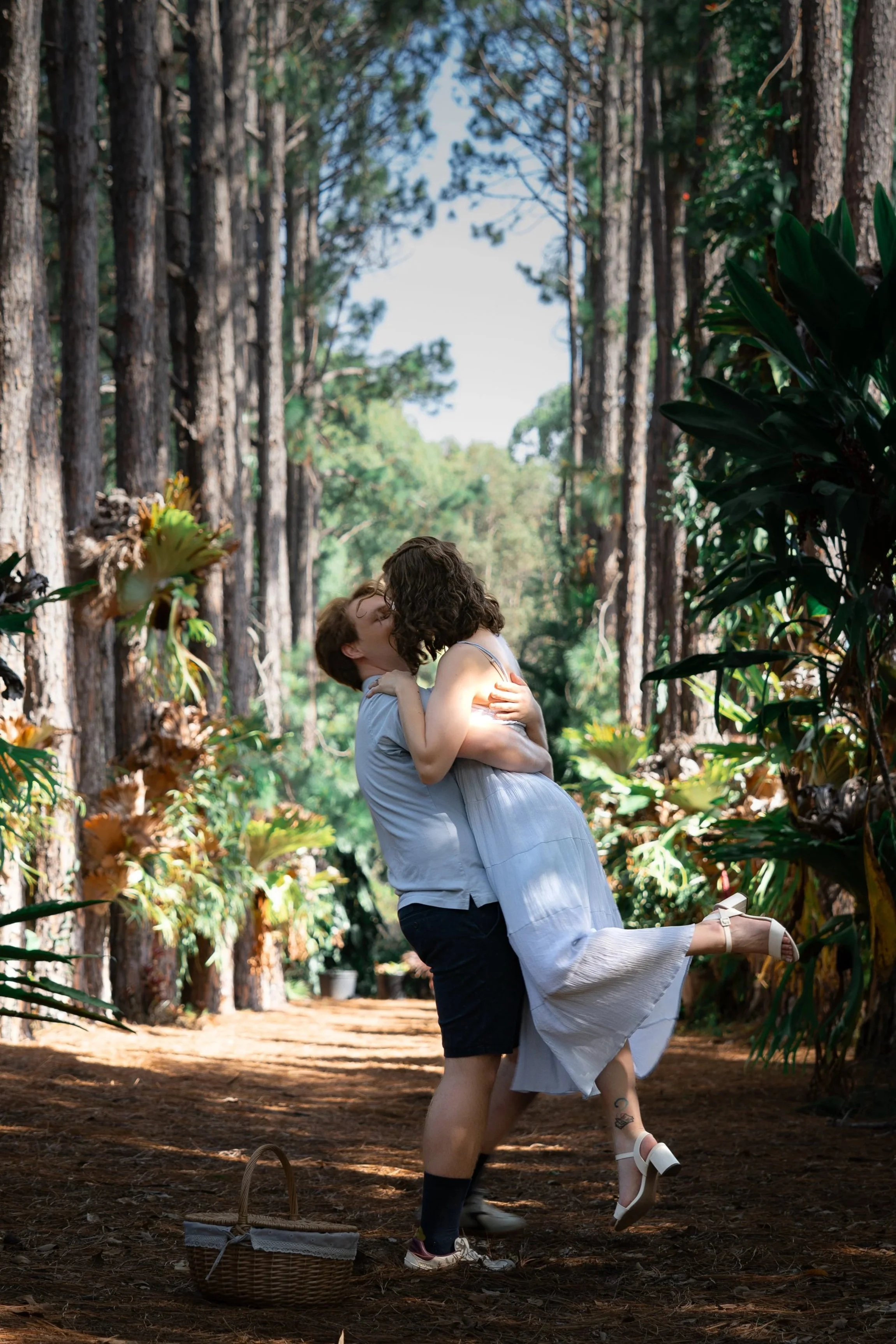 A couple sharing a kiss in a forested area, with the woman lifted by the man, surrounded by tall trees and lush greenery.
