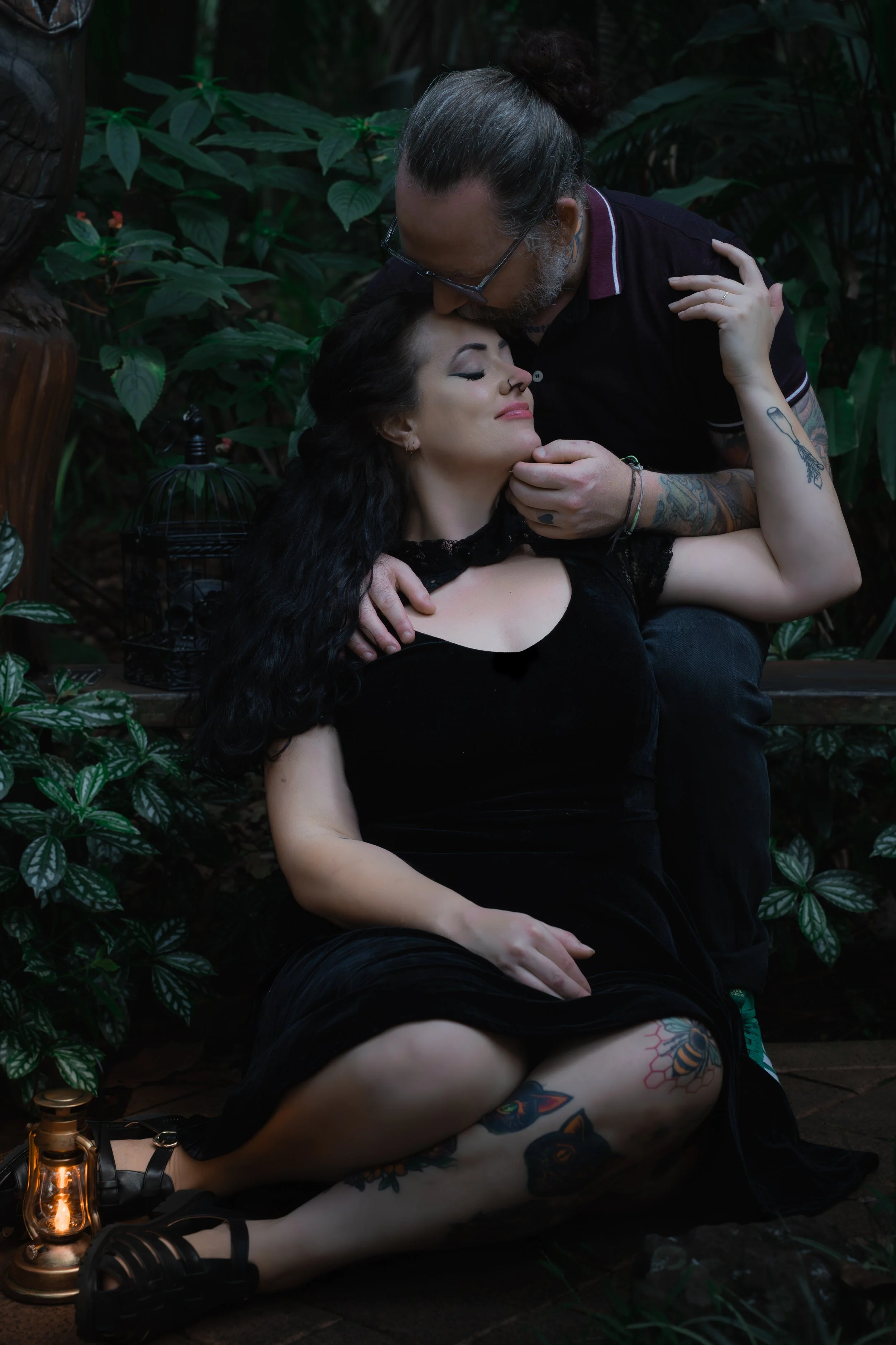 A man with long hair and tattoos gently kisses a woman with long dark hair and tattoos on her legs, sitting on the ground surrounded by green plants, while holding her head and shoulder. She is wearing a black dress and the scene is softly lit with a