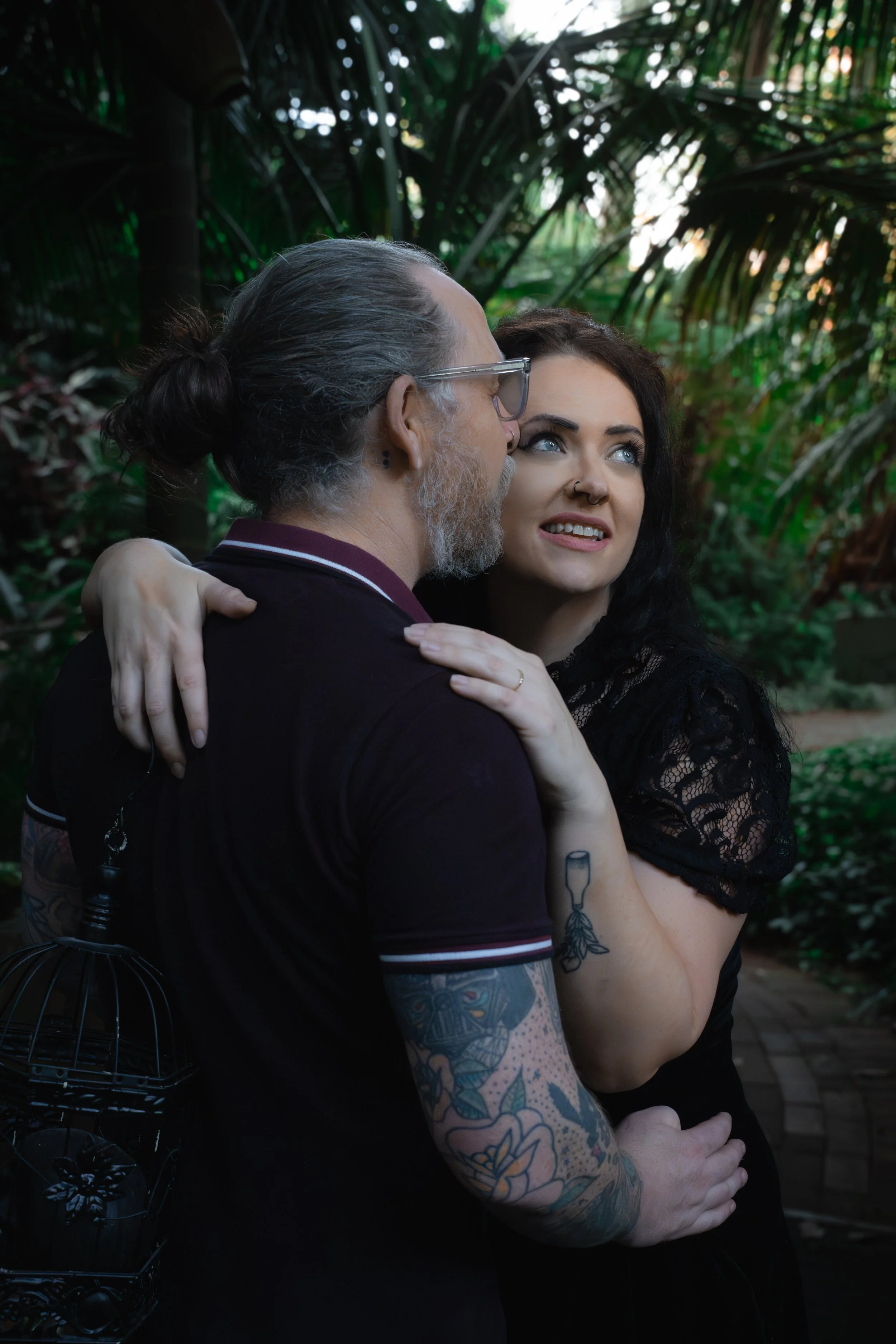 A man with long gray hair tied back, glasses, and tattoos on his arm embraces a woman with dark, curly hair, a nose piercing, and a tattoo on her arm, in a lush, green outdoor setting.
