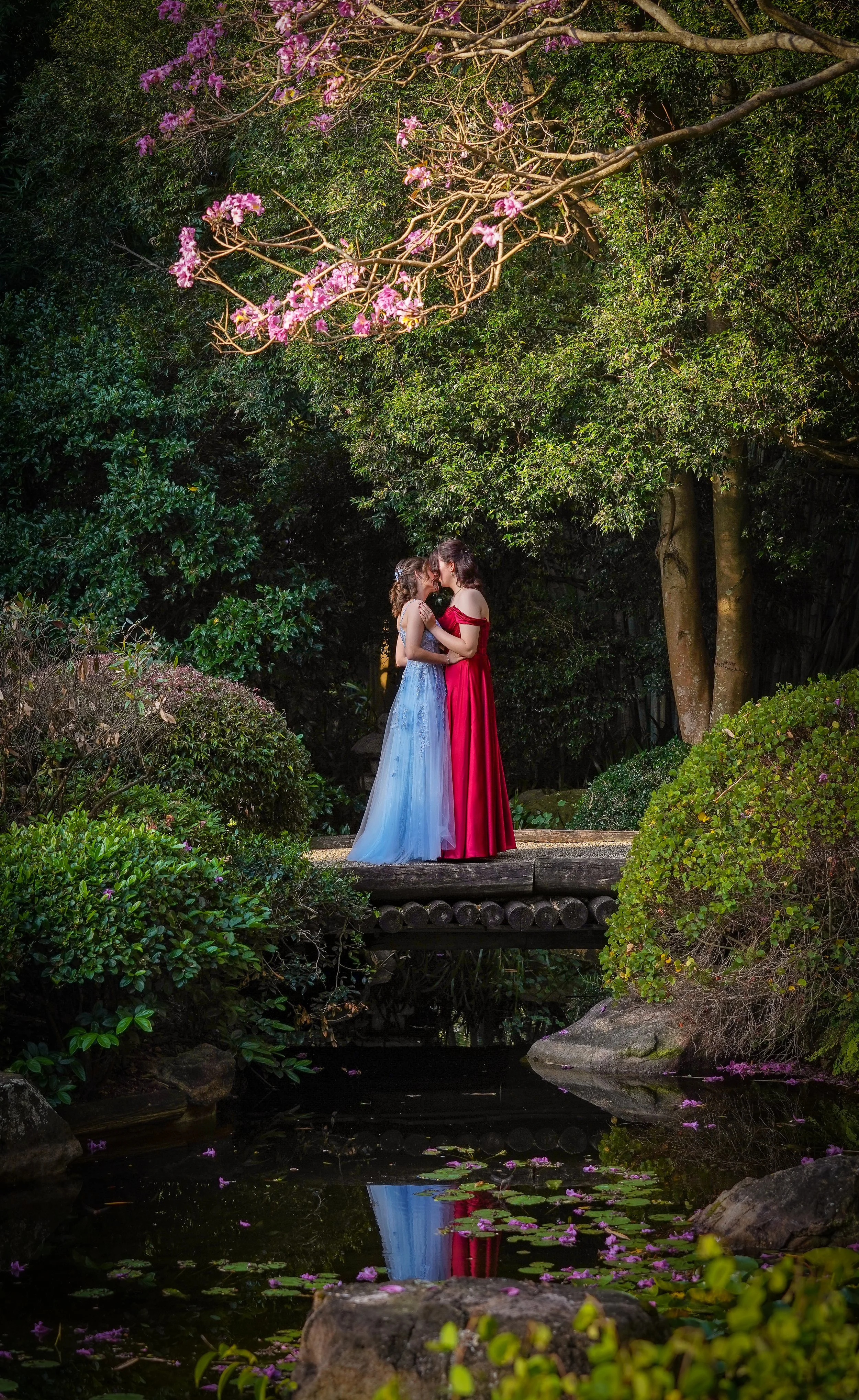 Two women in elegant dresses share a kiss on a small wooden bridge in a lush garden with colorful flowers and greenery. Their reflection is visible in the water below.