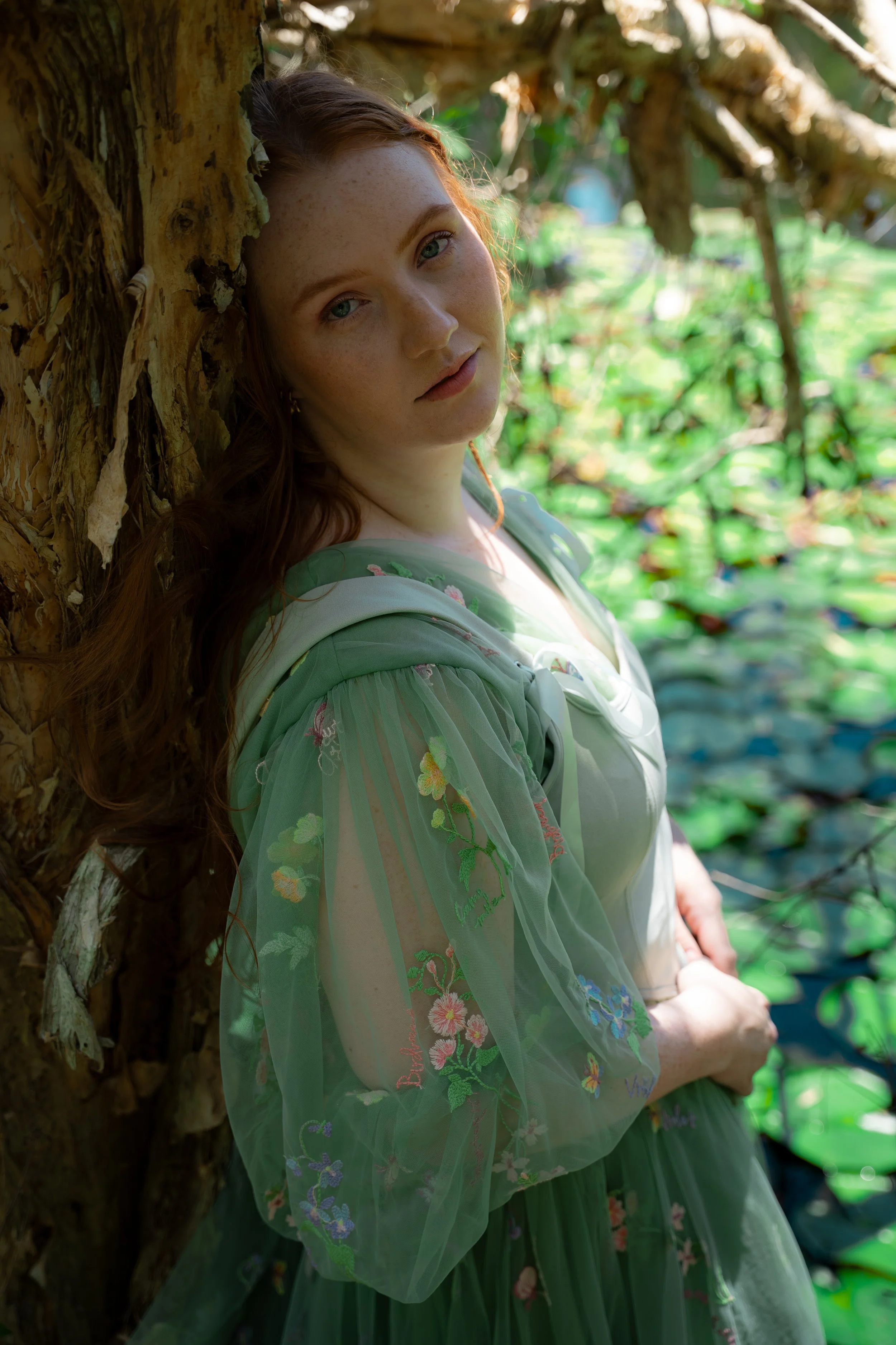 A woman with red hair and freckles leaning against a tree trunk in a lush, green outdoor setting, wearing a sheer, embroidered green dress.