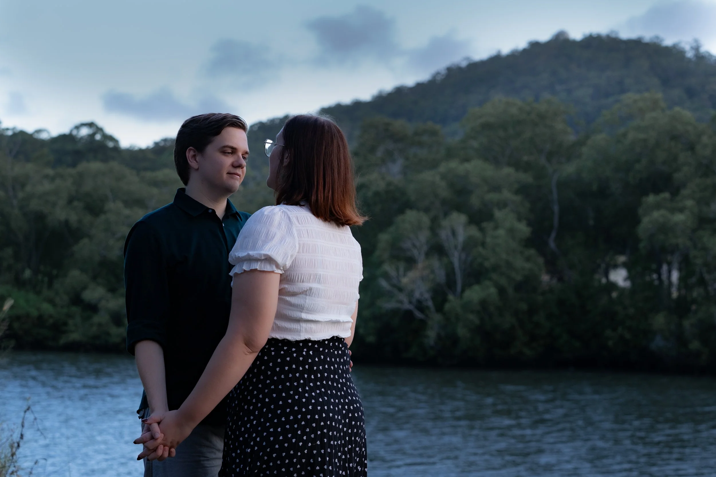 A young man and woman face each other holding hands outdoors near a body of water, with trees and hills in the background, during dusk or early evening.
