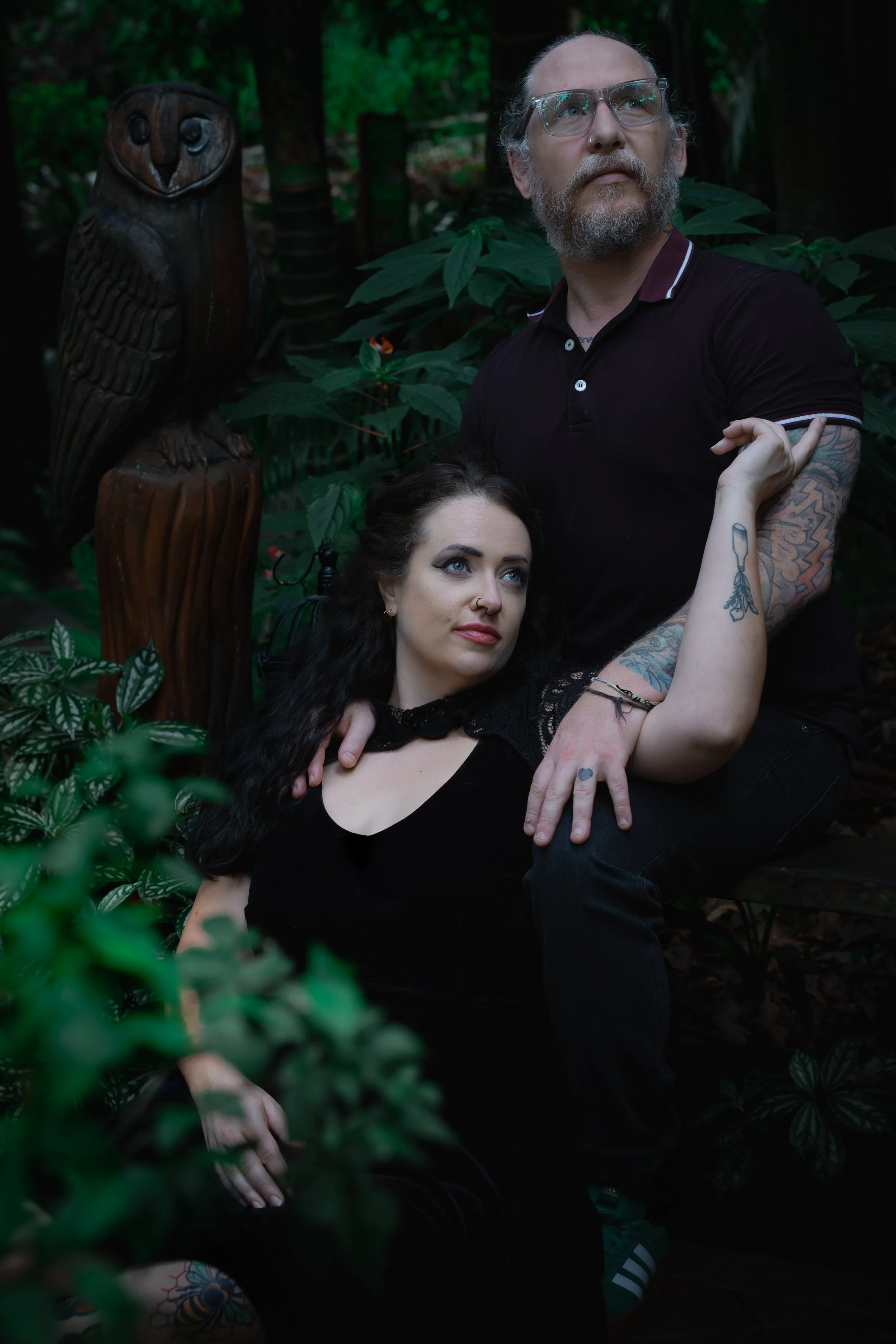 A man and a woman sit in a lush forest setting. The man is wearing glasses and a black polo shirt with tattoos on his arms, looking upwards. The woman, dressed in black with dark hair, rests her head on the man’s lap and gazes at him. There is a carv
