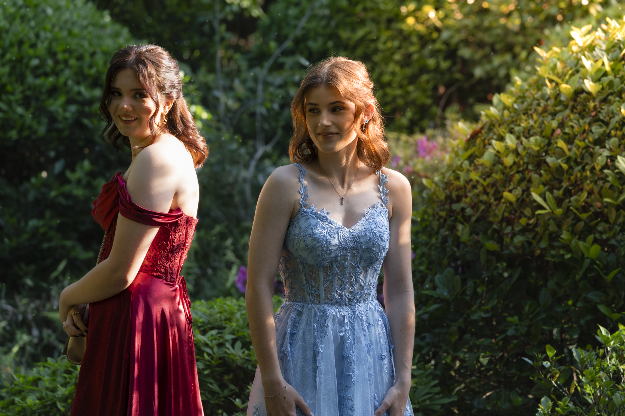 Two young women in elegant dresses standing outdoors in a garden, with lush green foliage and pink flowers in the background, illuminated by sunlight.