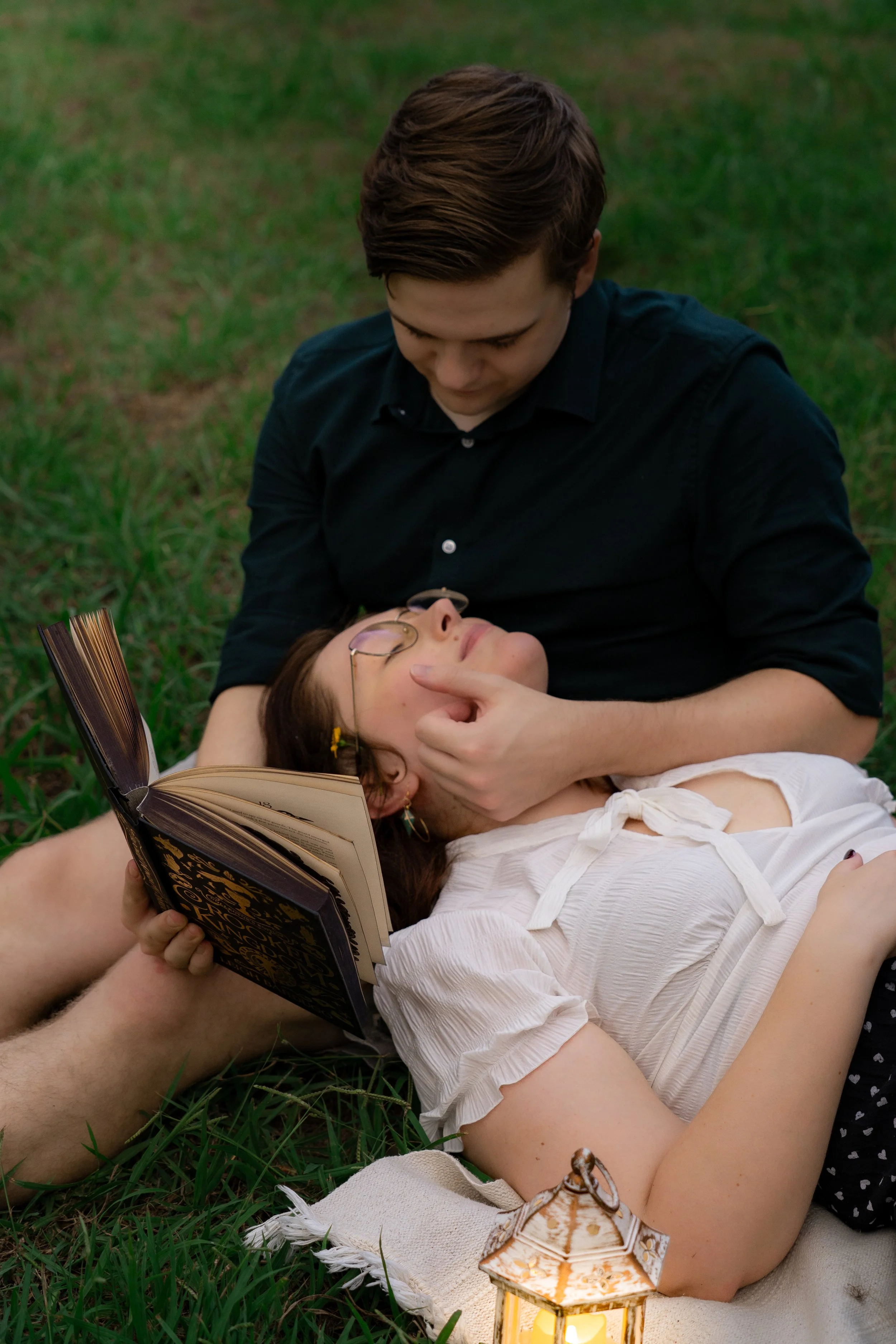 A young man with dark hair wears a black shirt and looks down at a young woman with glasses and brown hair, who is lying on her back in a white blouse with ribbons. The woman wears earrings and is holding an open book. They are outdoors on grass, wit