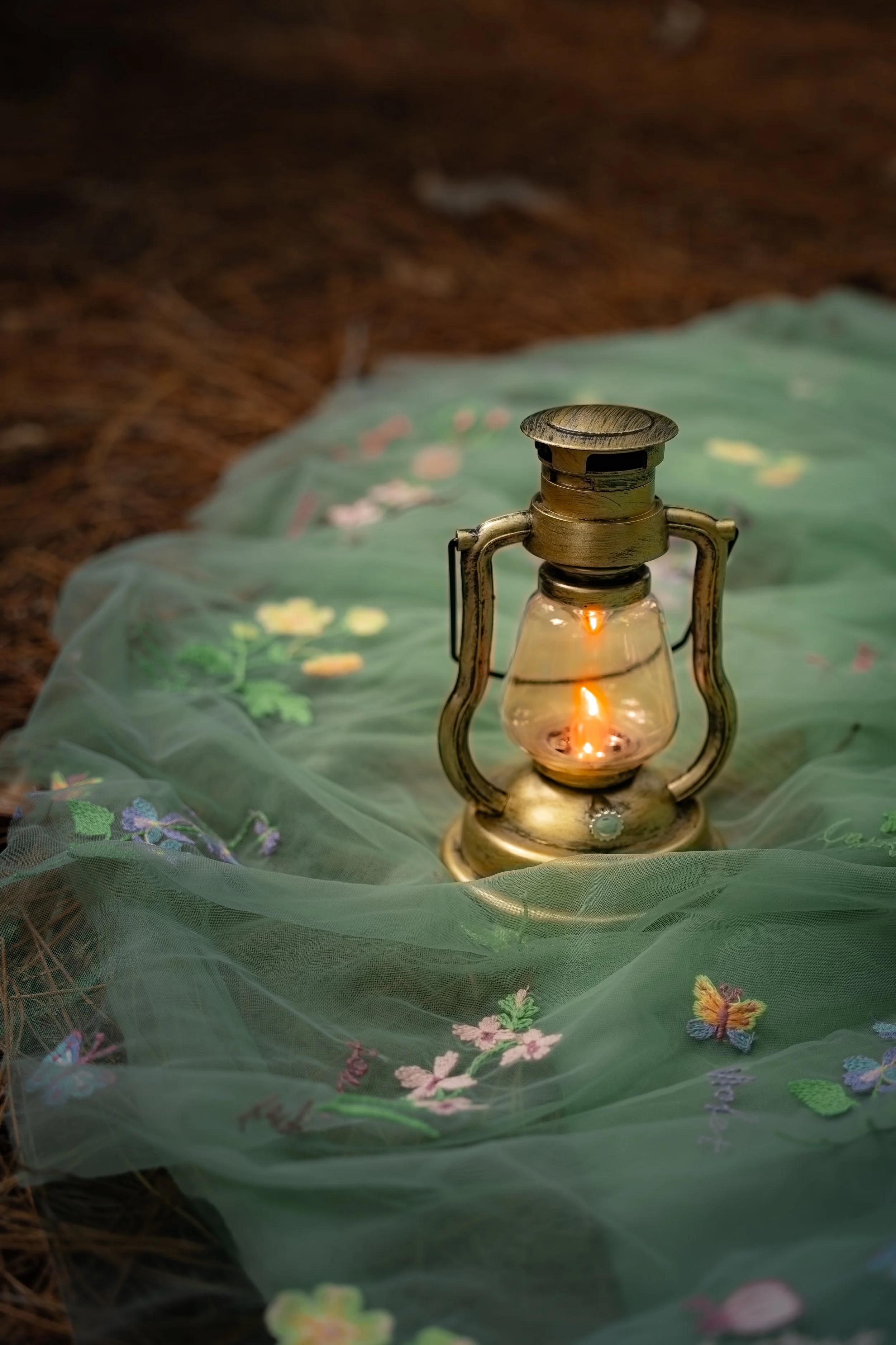 Vintage lantern with lit candle inside, placed on green embroidered fabric with floral and butterfly designs, on a forest floor.