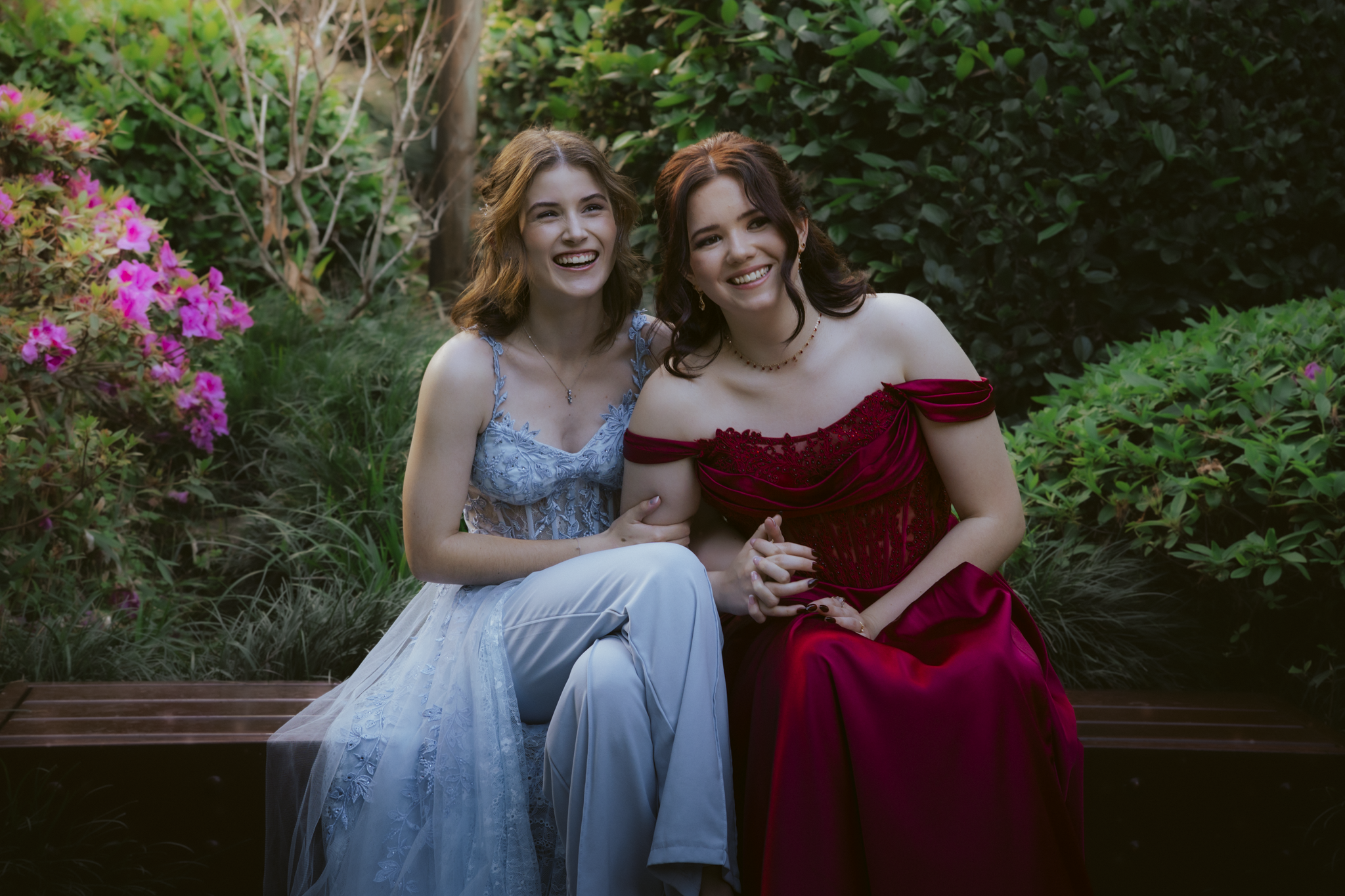 Two young women sitting on a wooden bench outdoors among green bushes and pink flowers, smiling and holding hands. One is wearing a light blue dress with lace details, and the other is wearing a red off-shoulder gown.