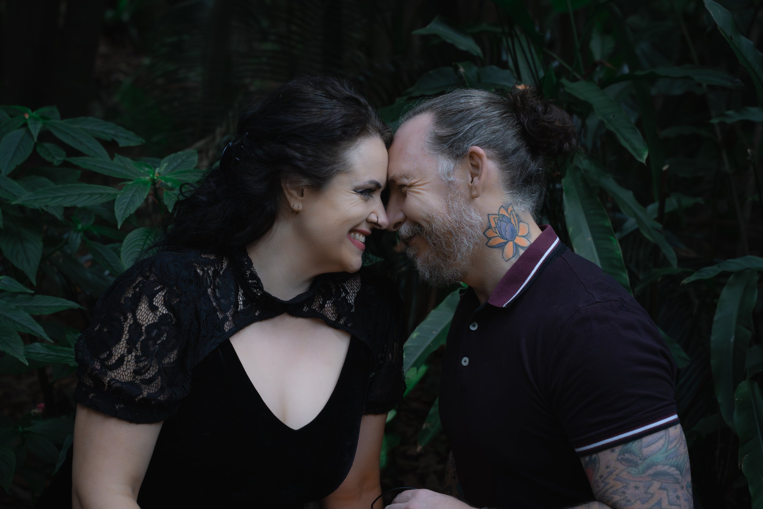 A man and woman touch foreheads and smile in a lush green outdoor setting.