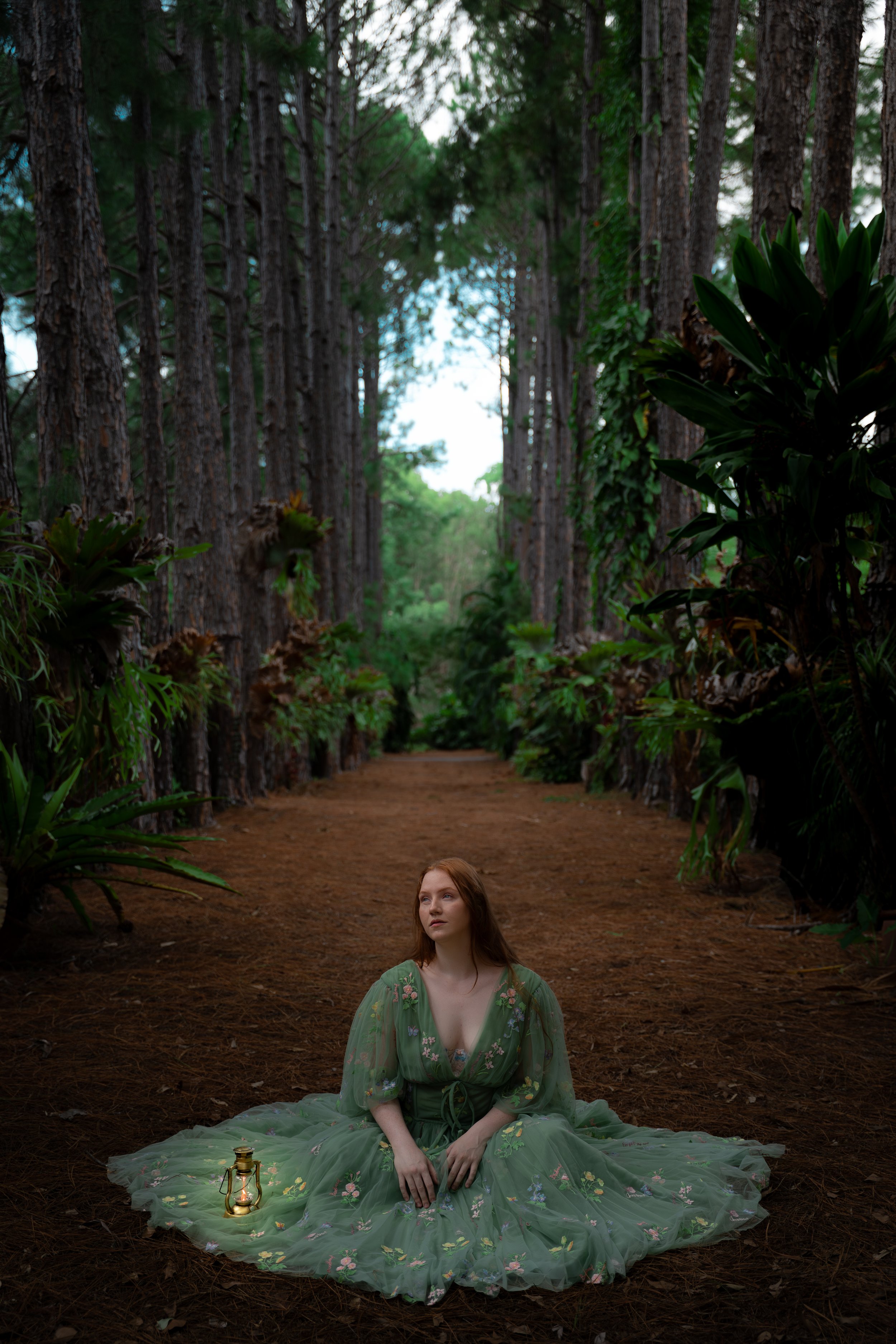 A woman with long red hair, dressed in a green embroidered gown, sitting cross-legged on a forest trail surrounded by tall trees and lush green foliage during daytime, with a lantern placed beside her.