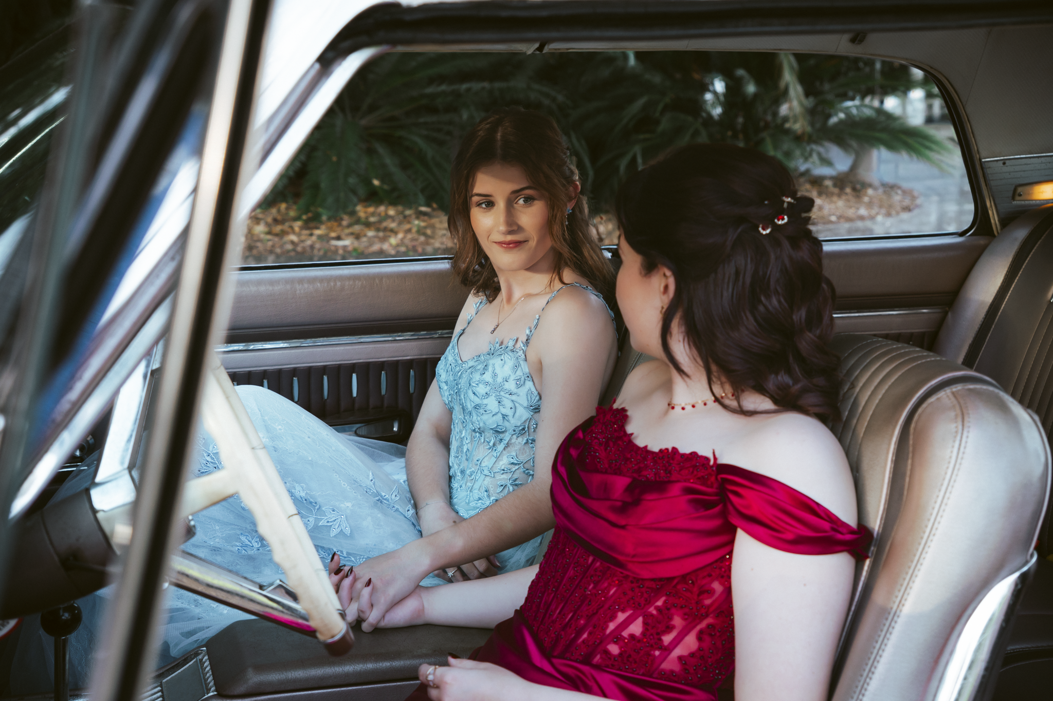 Two young women sitting in the front seats of a vintage car, holding hands with their fingers intertwined, dressed in colorful evening gowns with a background of trees and greenery.
