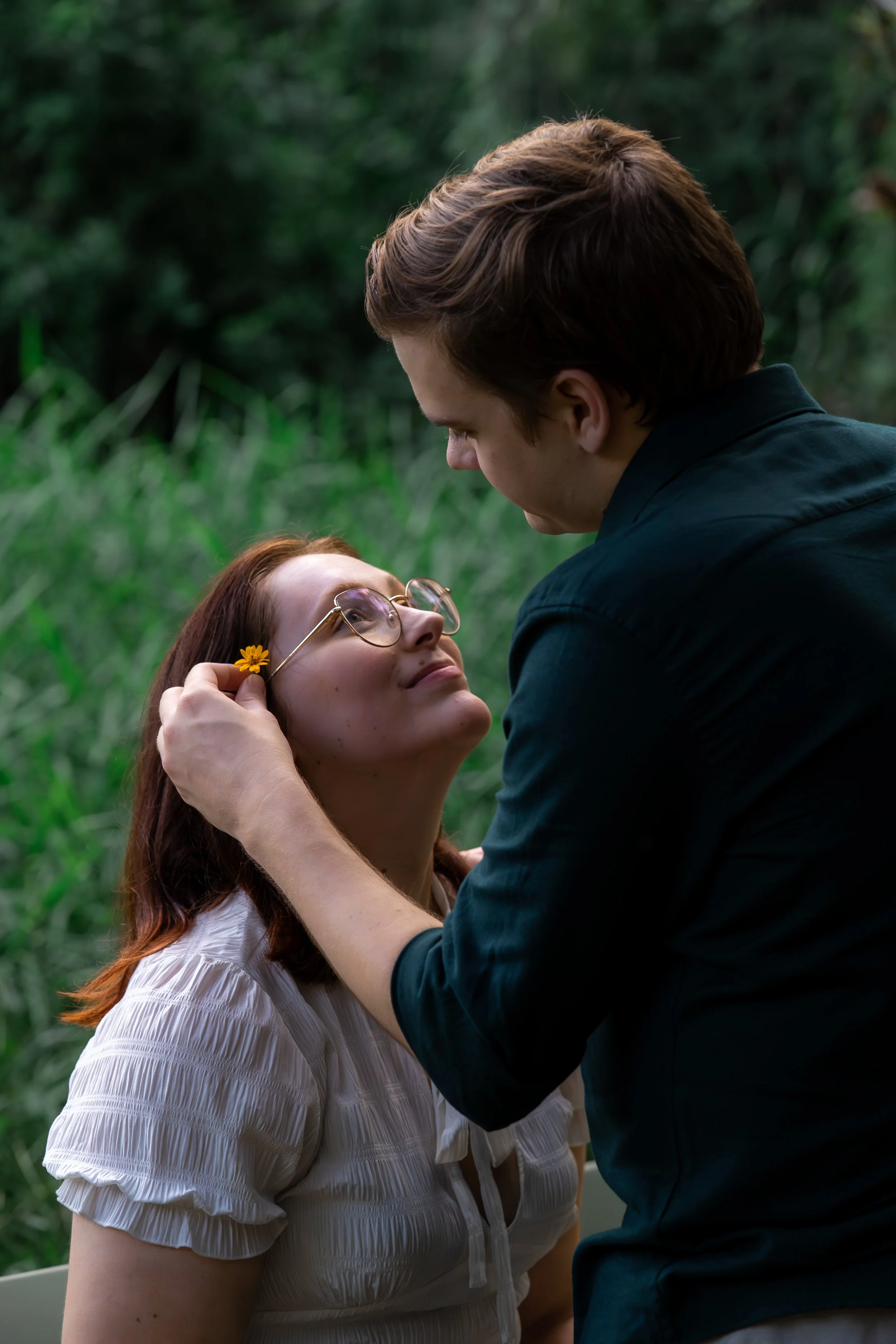 A man kneeling in front of a woman outdoors, holding her face gently with one hand, looking into her eyes lovingly. The woman is smiling softly, wearing glasses with a small flower tucked behind one ear. They are surrounded by greenery.
