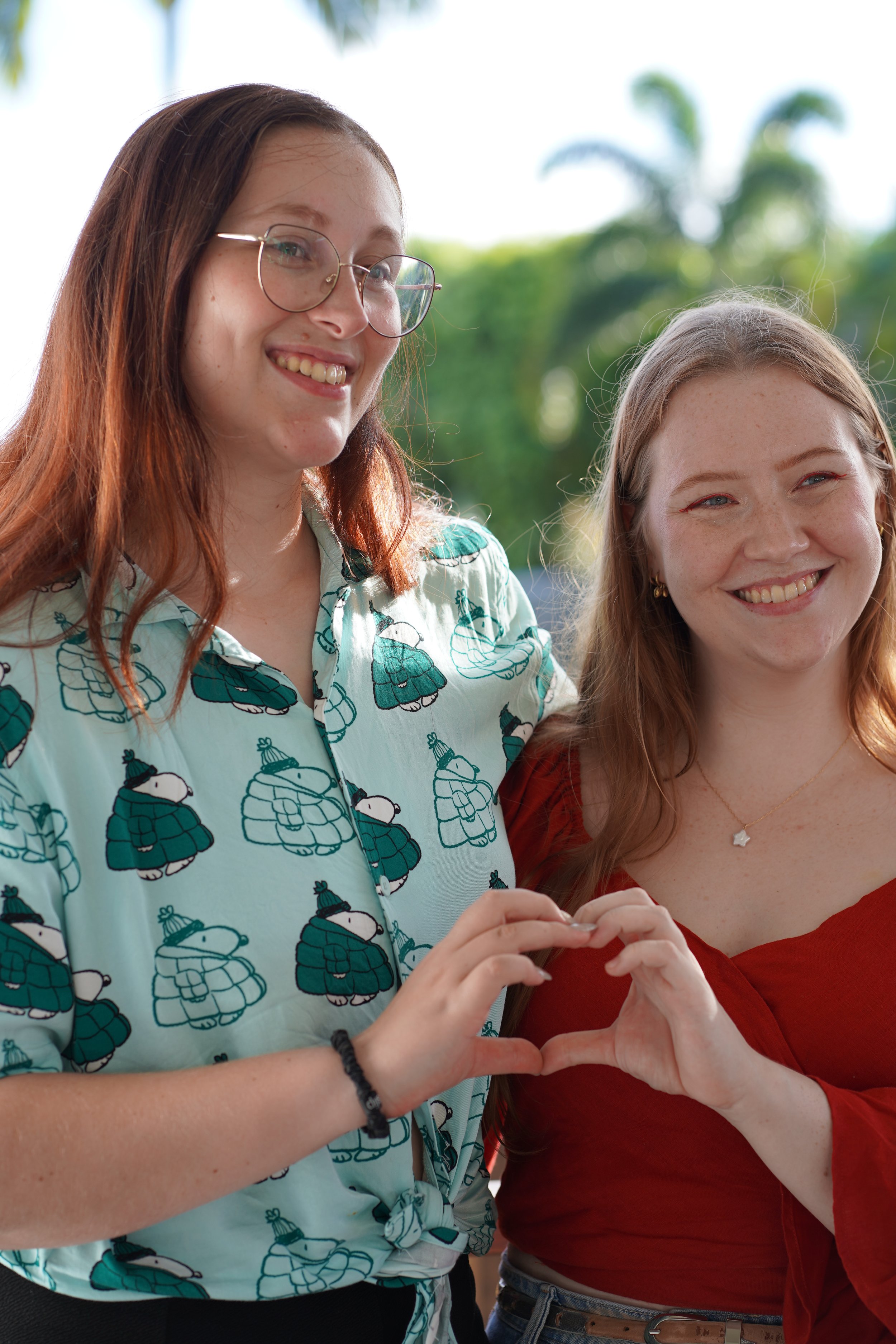 Two women smiling and making a heart shape with their hands outdoors.