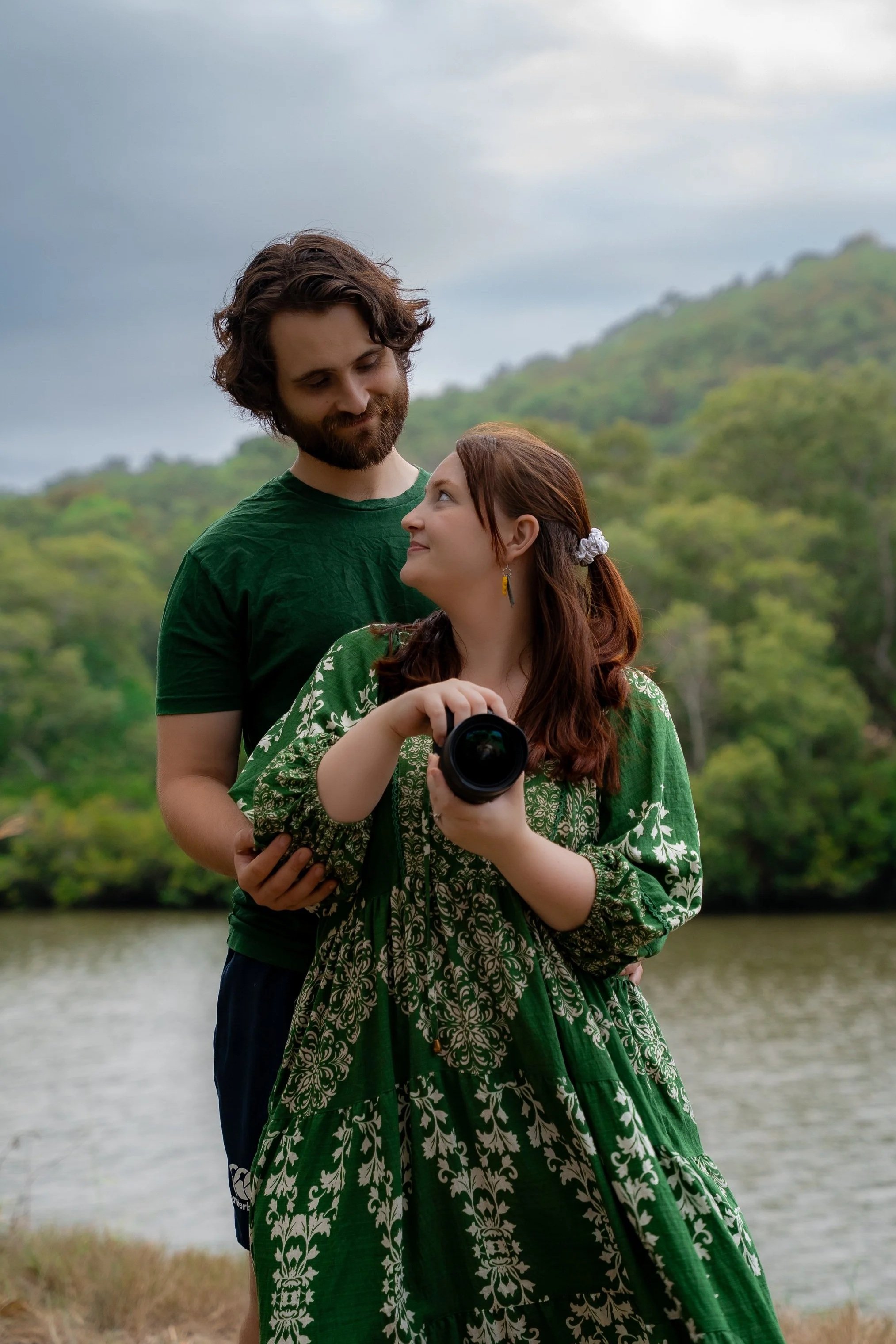A man and woman standing close by a lake with greenery and hills in the background. The woman is holding a camera and looking up at the man, who is looking down at her. The woman has red hair with a hair clip and is wearing a green patterned dress. The man has dark hair and a beard, wearing a green T-shirt.