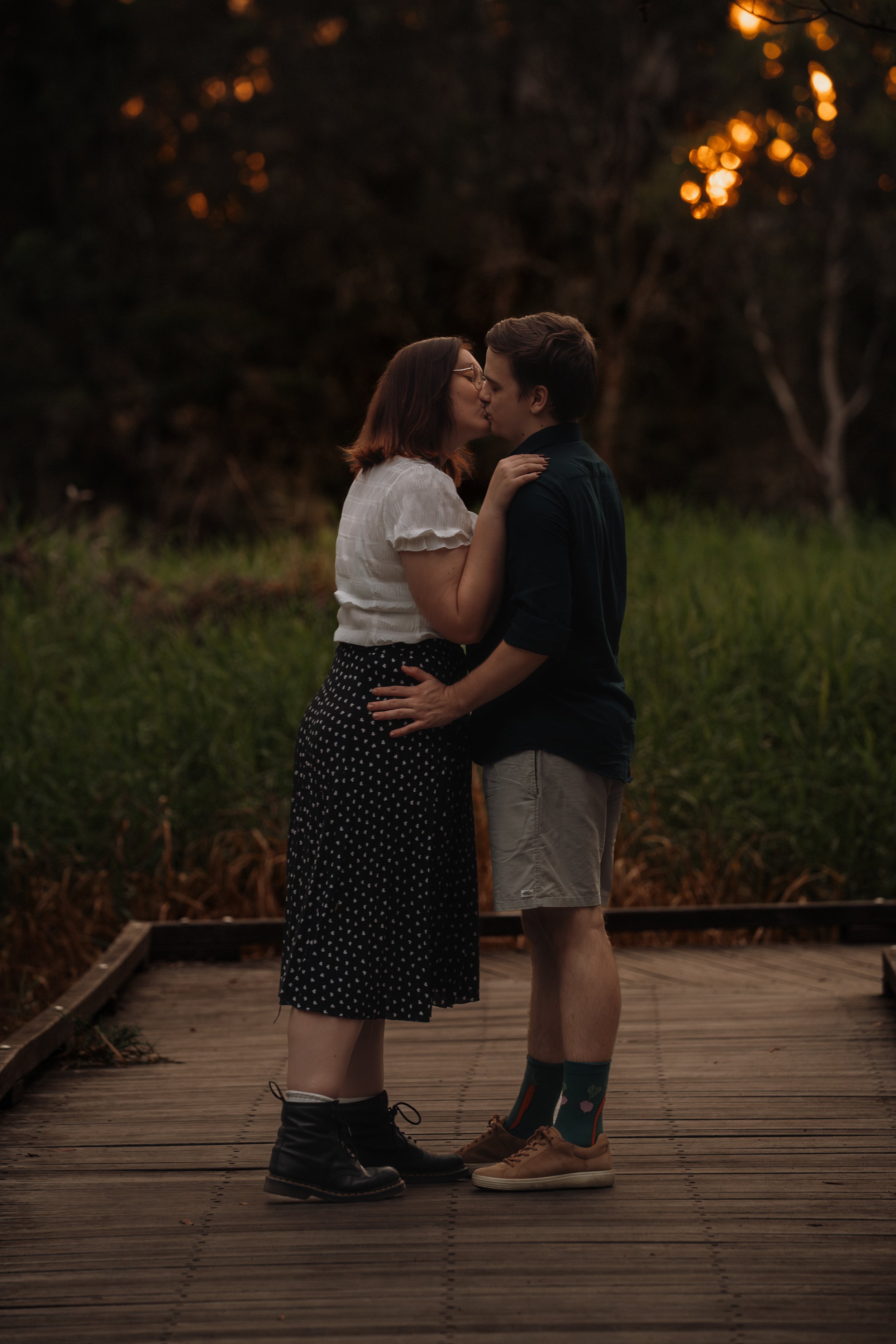 A couple kissing on a wooden walkway at sunset, surrounded by trees and greenery.