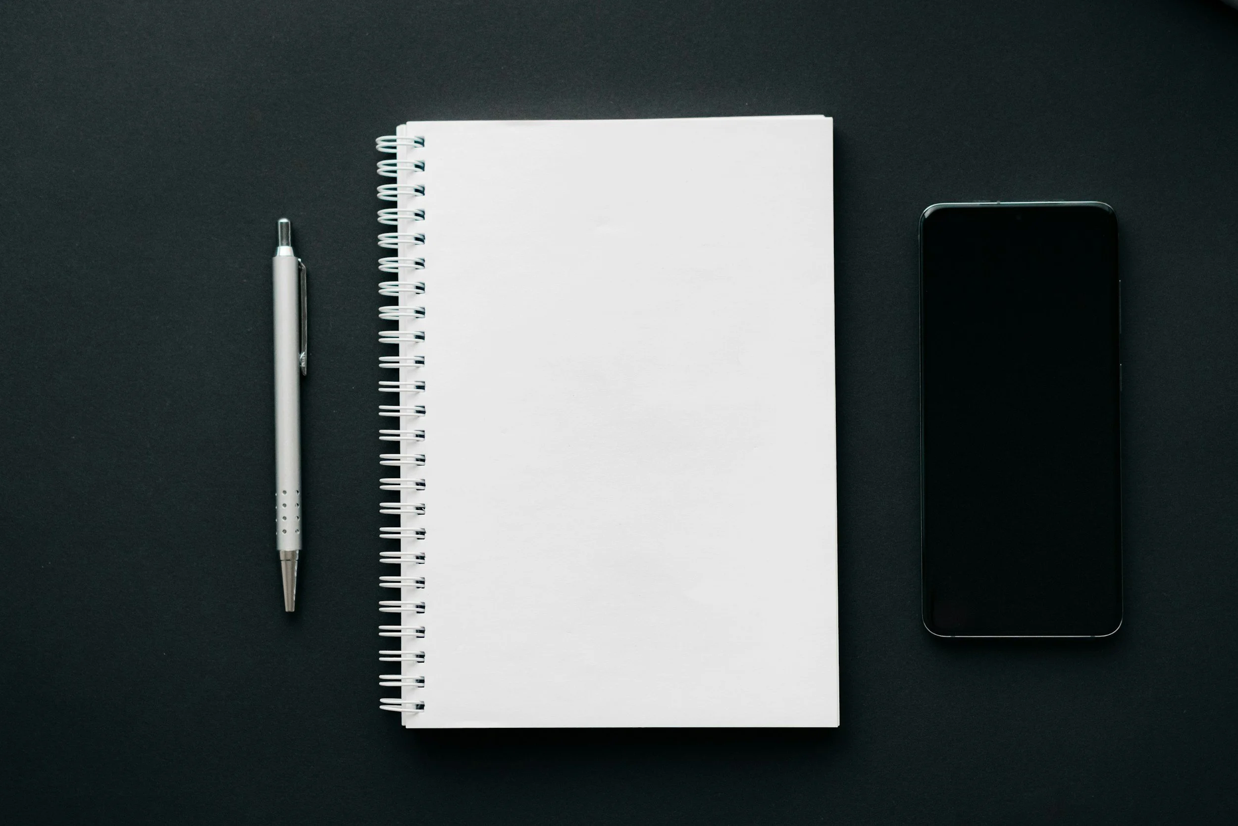 A flat lay photo of a silver pen, a spiral notebook with blank white pages, and a black smartphone on a black surface.