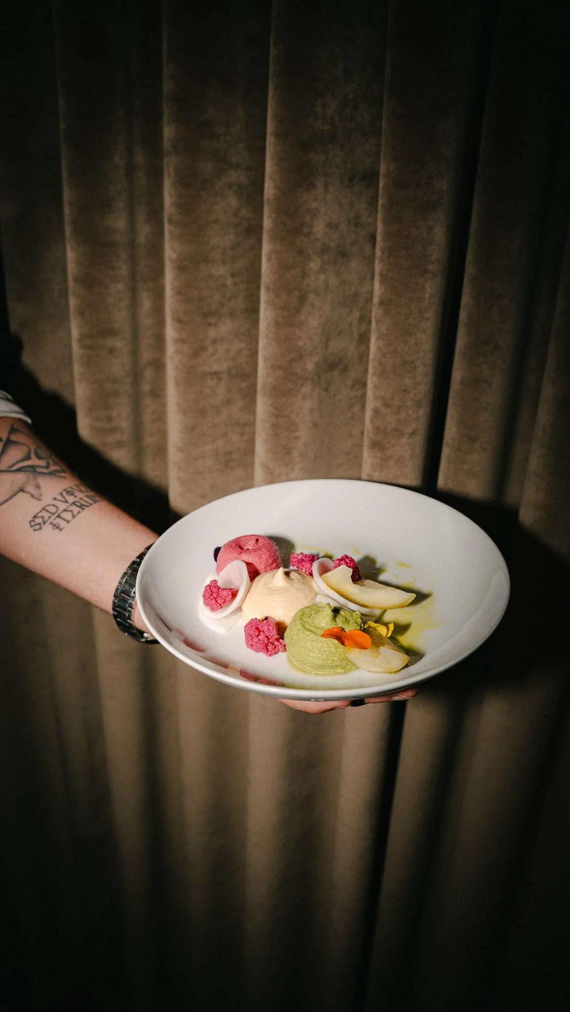 Person holding a white plate with assorted colorful dessert scoops and garnishes, standing in front of brown curtains.