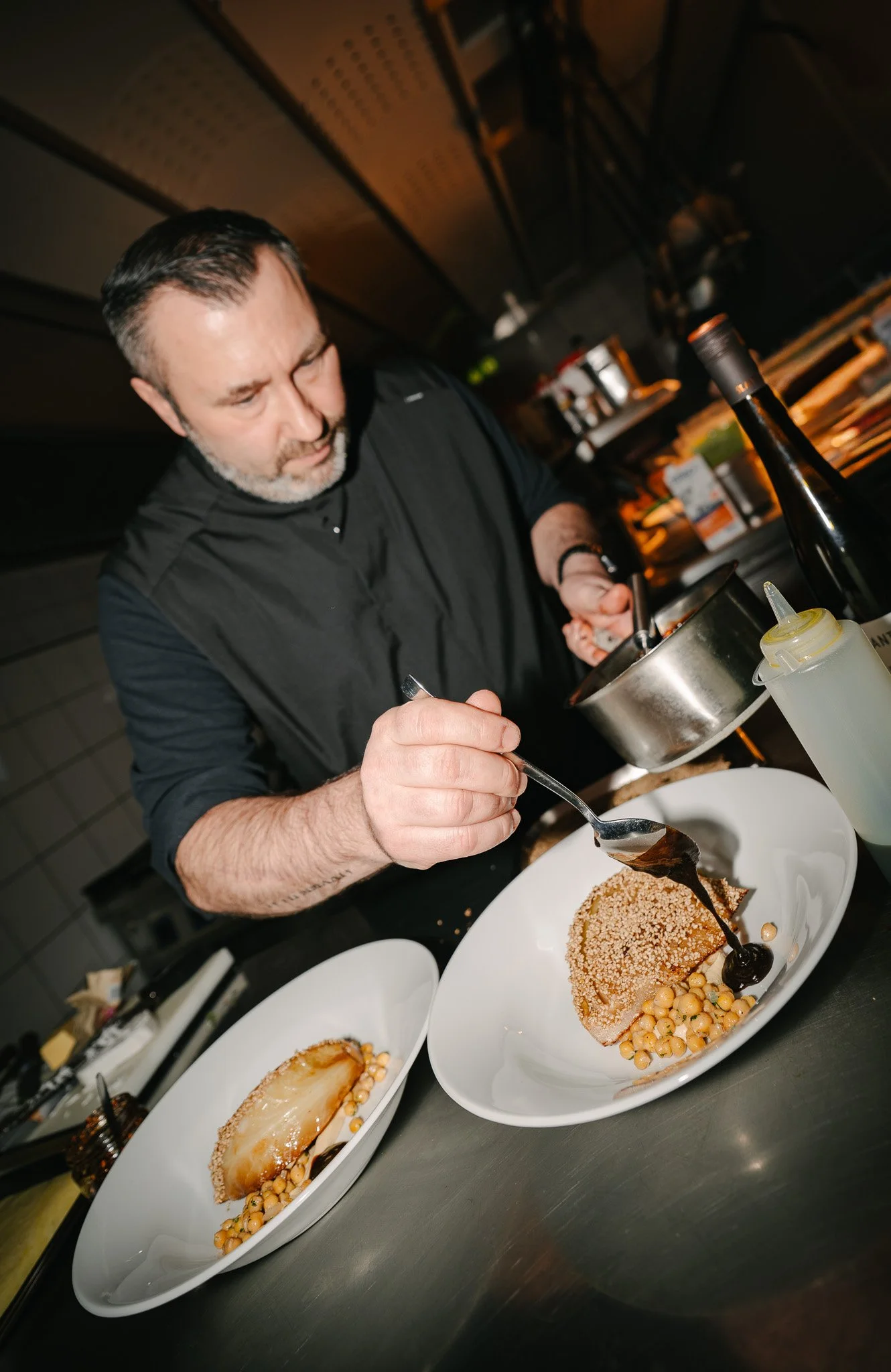 A chef is plating a dessert with a sauce in a professional kitchen, with two plates of the same dessert on the counter.