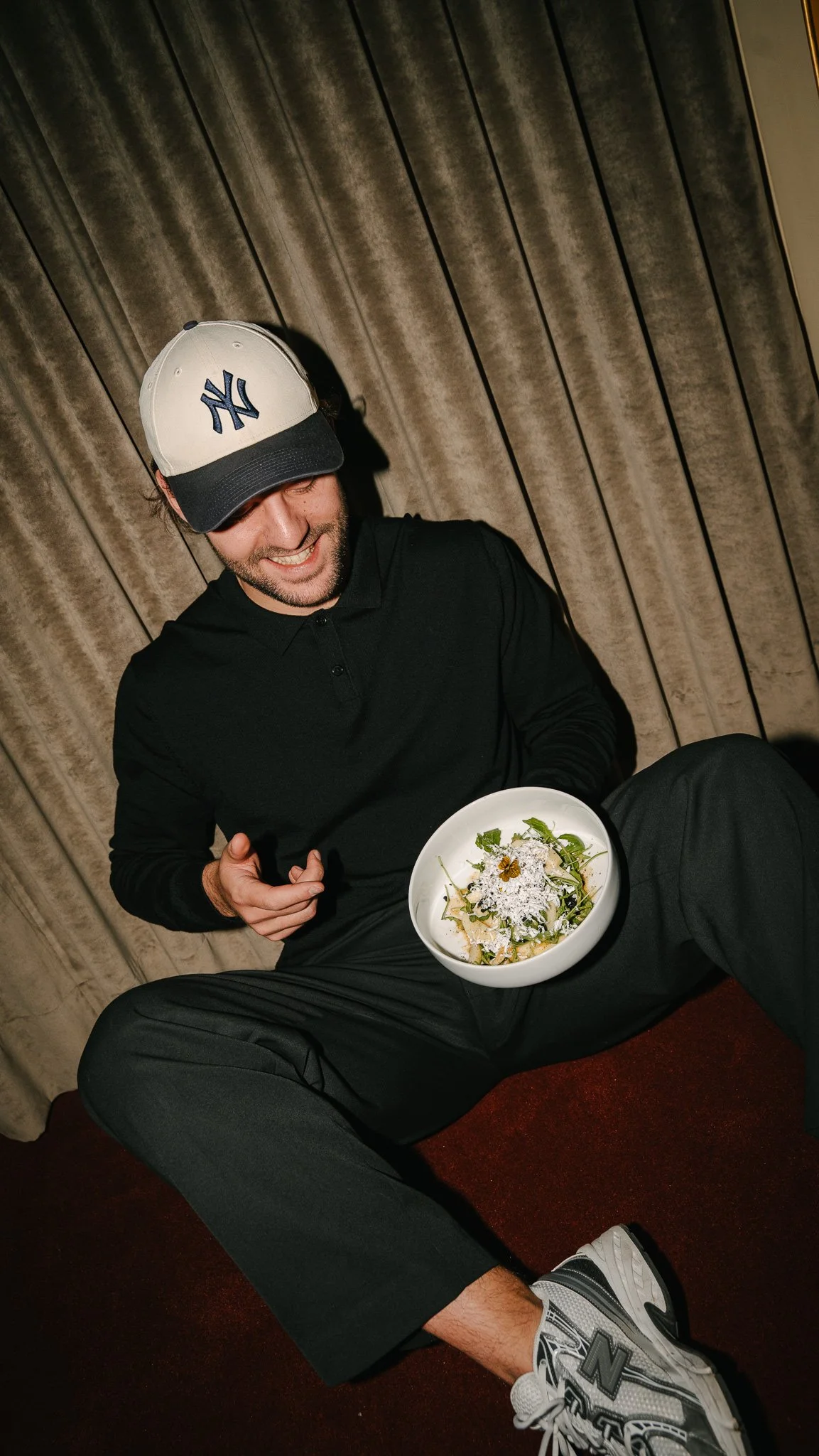 A man smiling and sitting with a bowl of salad, wearing a baseball cap and casual clothing, against a curtain background.