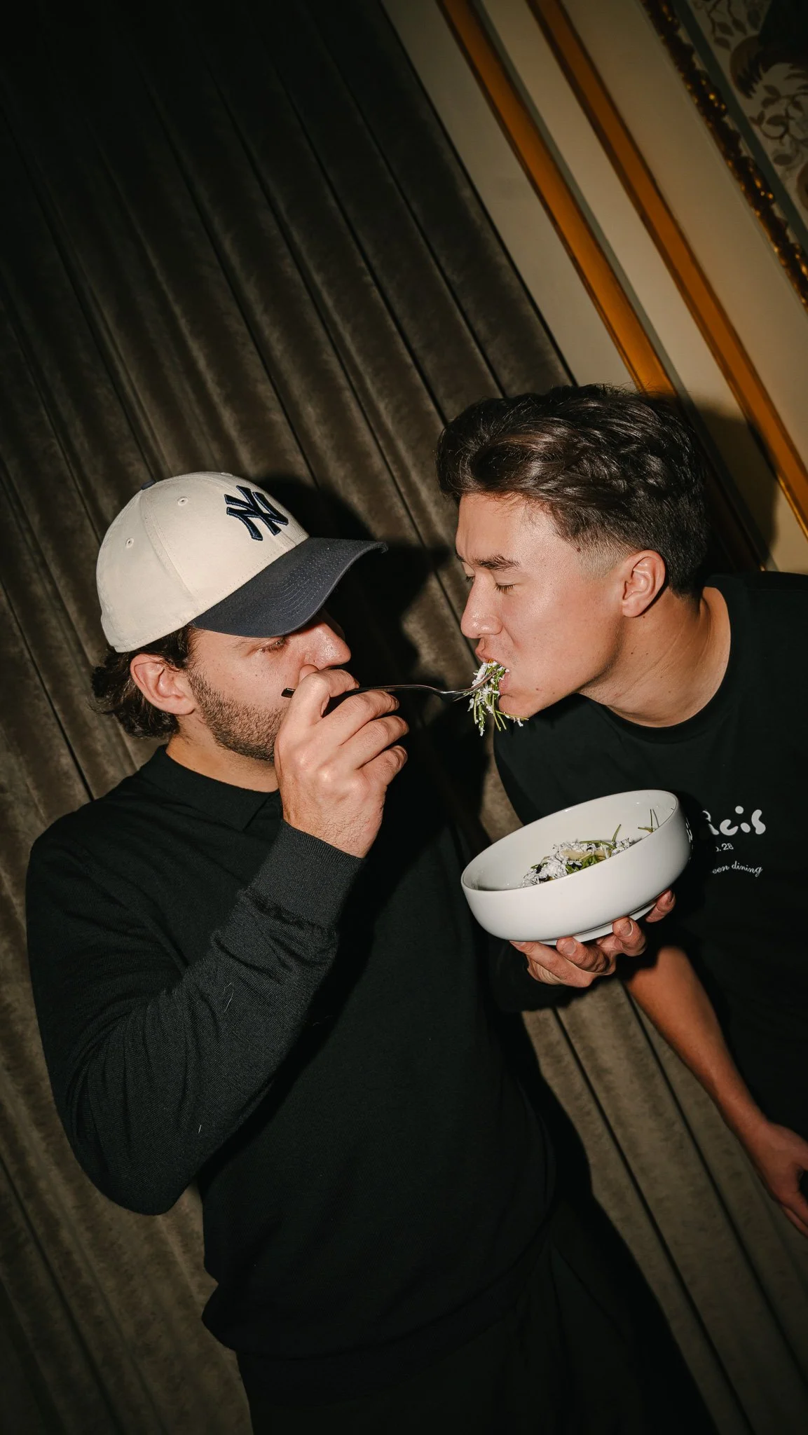 Two men sharing a bowl of salad, with one feeding the salad to the other at an indoor setting with dark curtains.