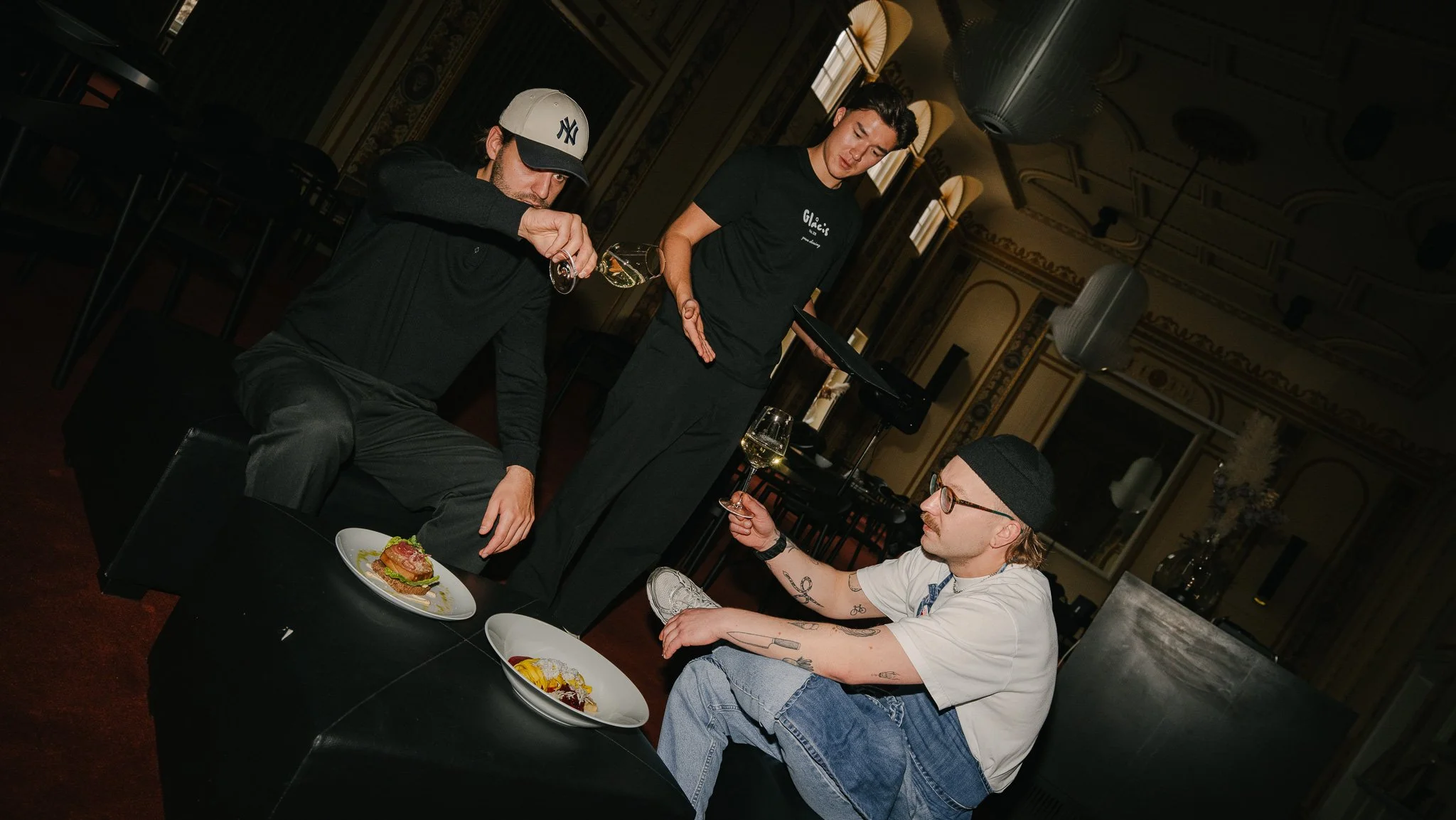 Two men sitting on black chairs with plates of food in front of them, and a woman standing between them pouring wine into one man's glass in a dimly lit, ornate room.