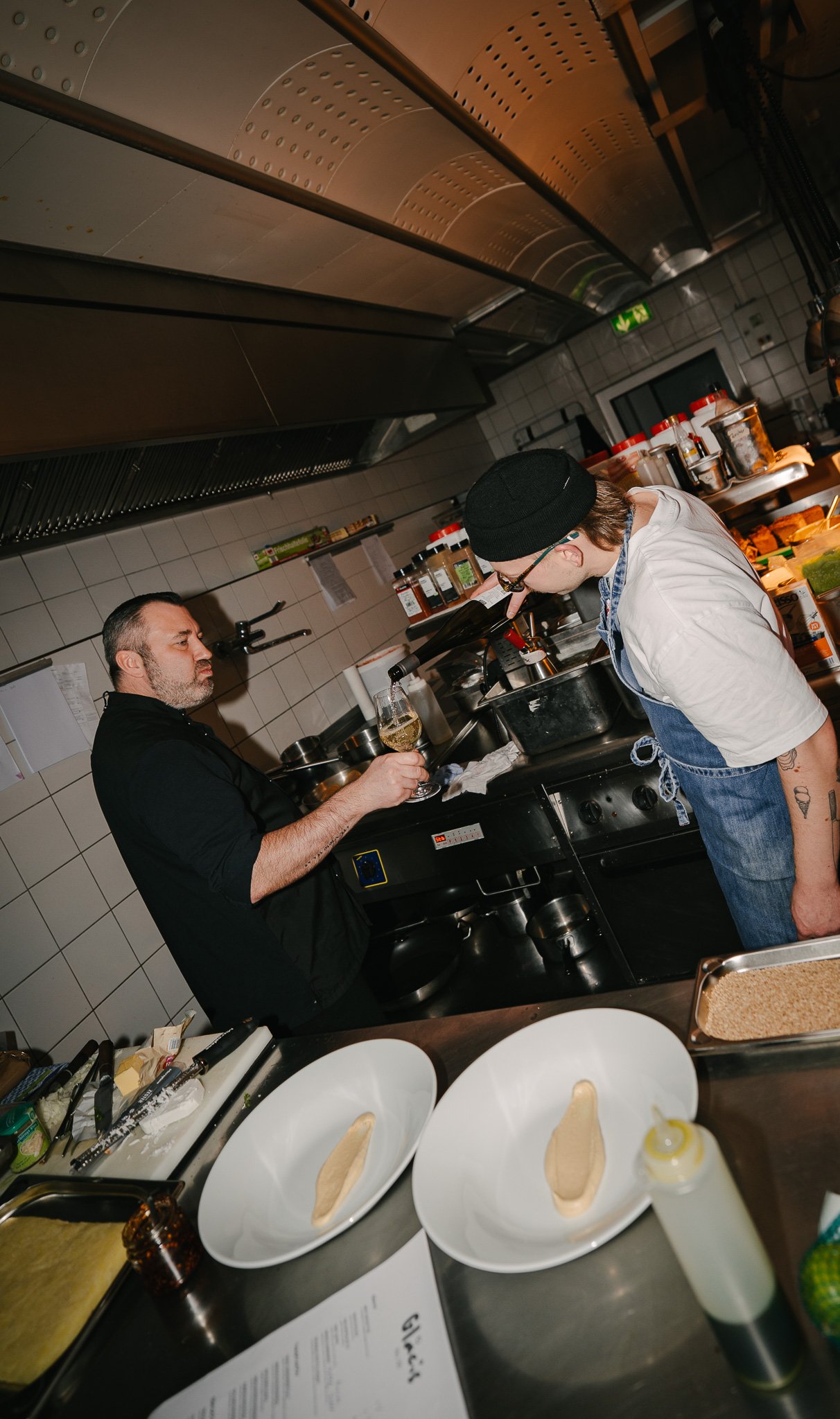 A chef and a man in a kitchen, with the man holding a glass of white wine and looking at the chef, who is leaning over the counter. The kitchen has various bottles and utensils on the counter and shelves.