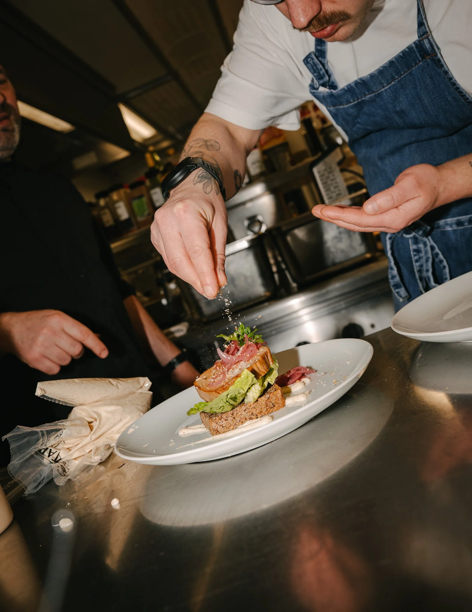 A chef is seasoning a gourmet dish with salt, served on a white plate with various ingredients including lettuce, meat, and sauce, in a restaurant kitchen.