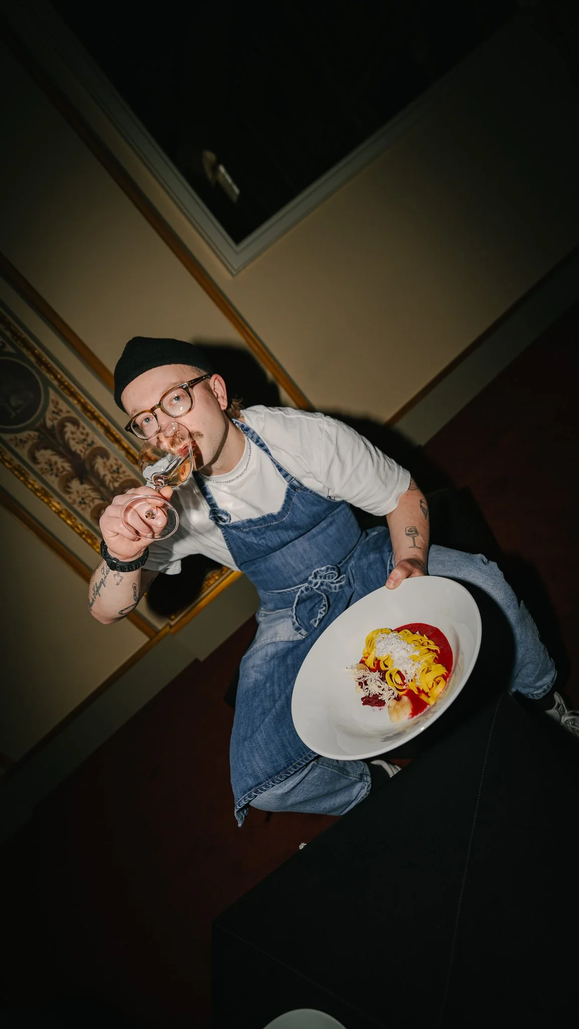 A man with glasses, tattoos, a black beanie, and a white shirt, wearing an apron, sitting and holding a fork with a glass of red wine, and a large plate of pasta with shredded cheese and sauce.