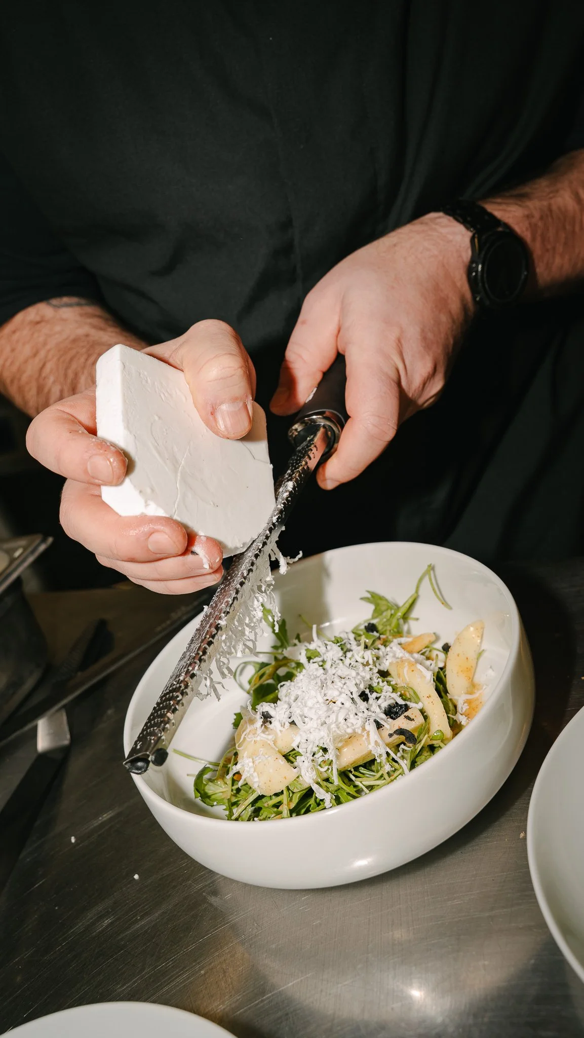 Person grating cheese over a bowl of salad with greens, sliced pears, and black olives.