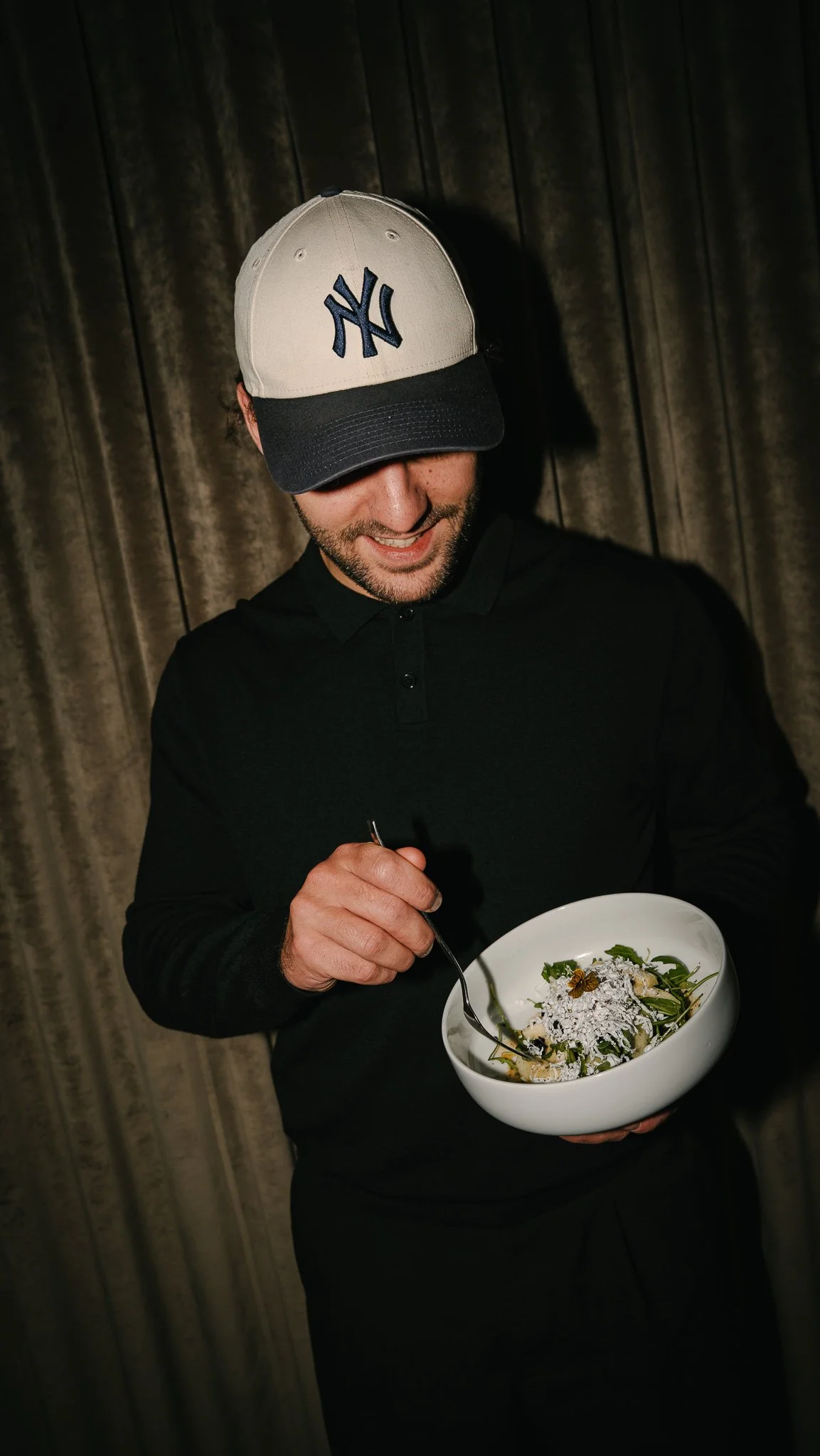 A man wearing a New York Yankees baseball cap, black shirt, and smiling while holding a bowl of salad with greens, cheese, and shredded toppings, standing in front of a dark curtain.