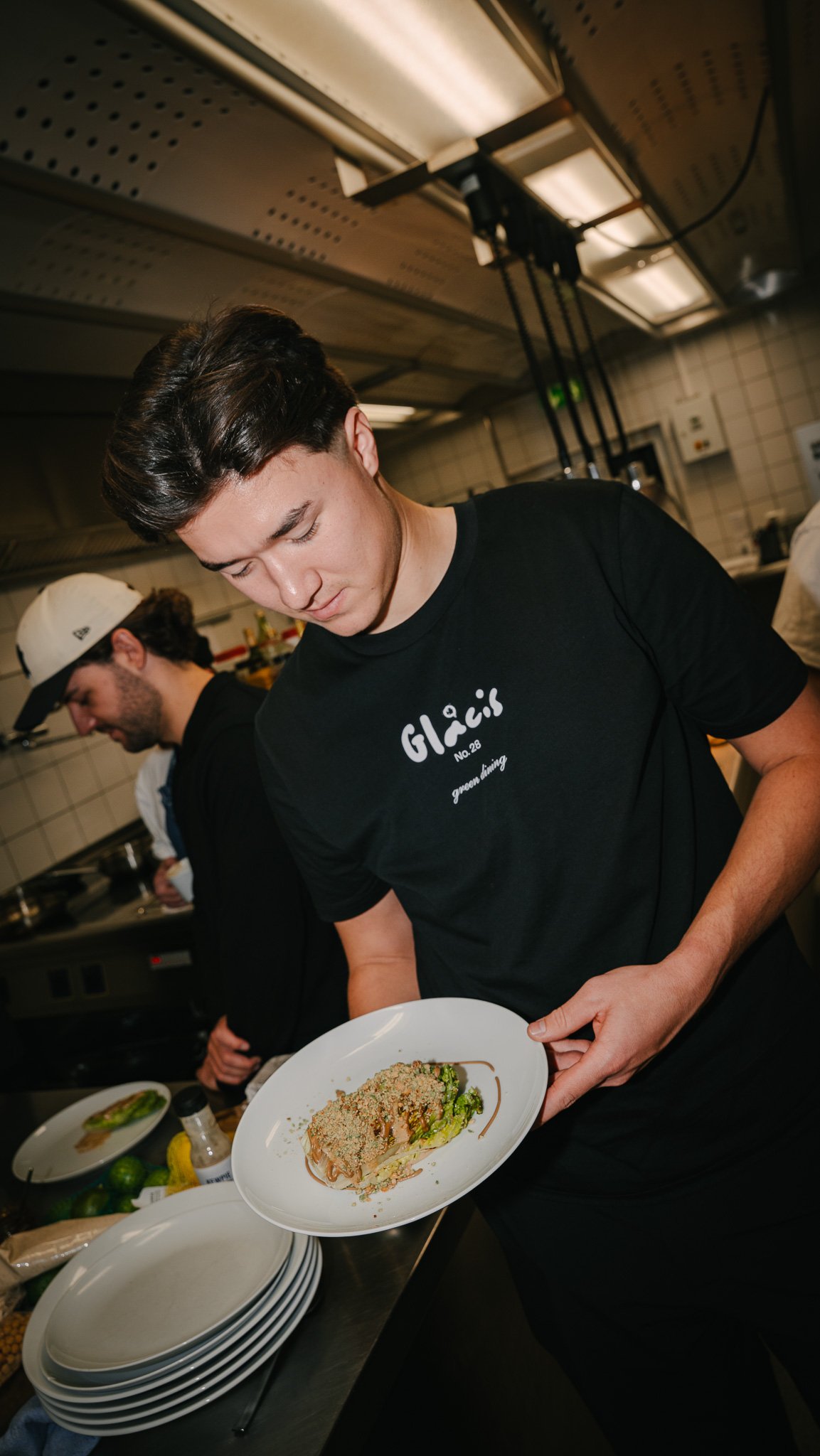 A young man in a black shirt with white text and a black apron holding a plated dish in a professional kitchen. Another person is visible in the background.