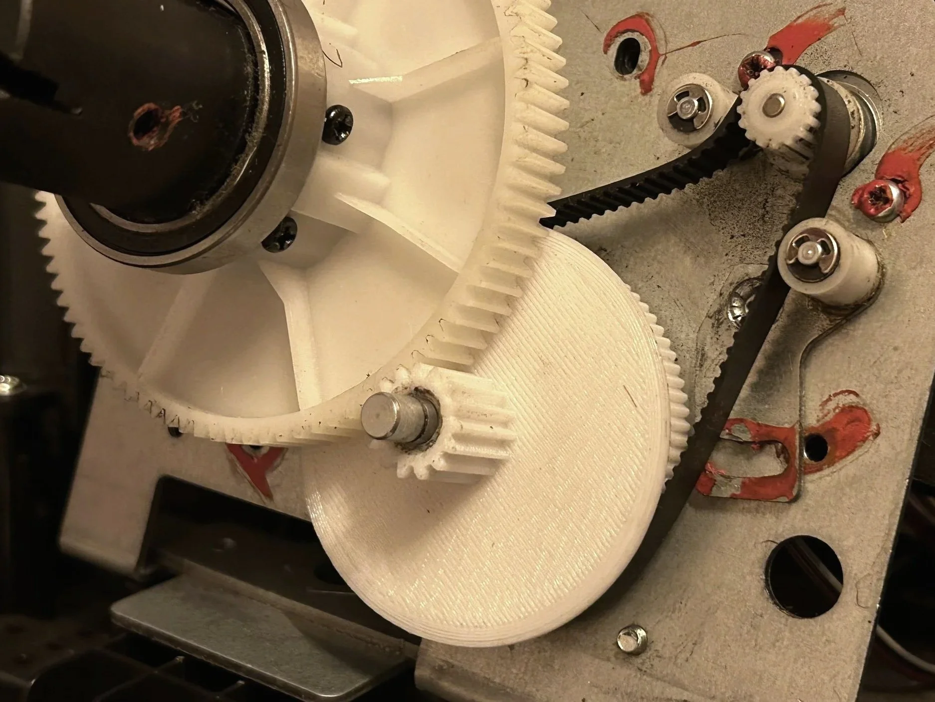Une vue de l'intérieur d'une machine avec des engrenages blancs, une ceinture noire et plusieurs composants métalliques, certains avec de la graisse rouge. La machine semble être en cours d'assemblage ou de maintenance.
