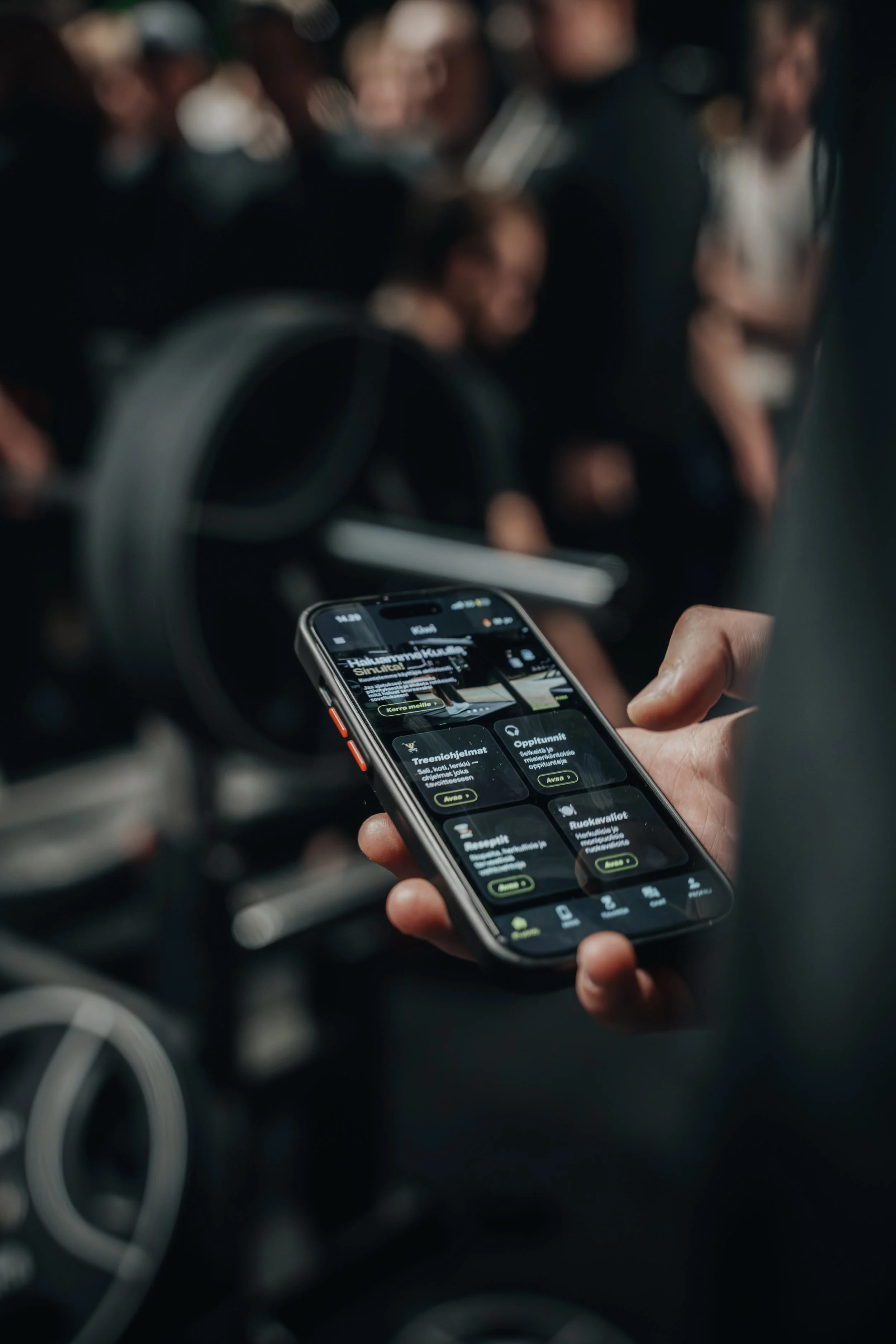Henkilö katsoo älypuhelintaan, jossa näkyy mobiilisovelluksen ekran, taustalla ihmisiä kokoontuneena