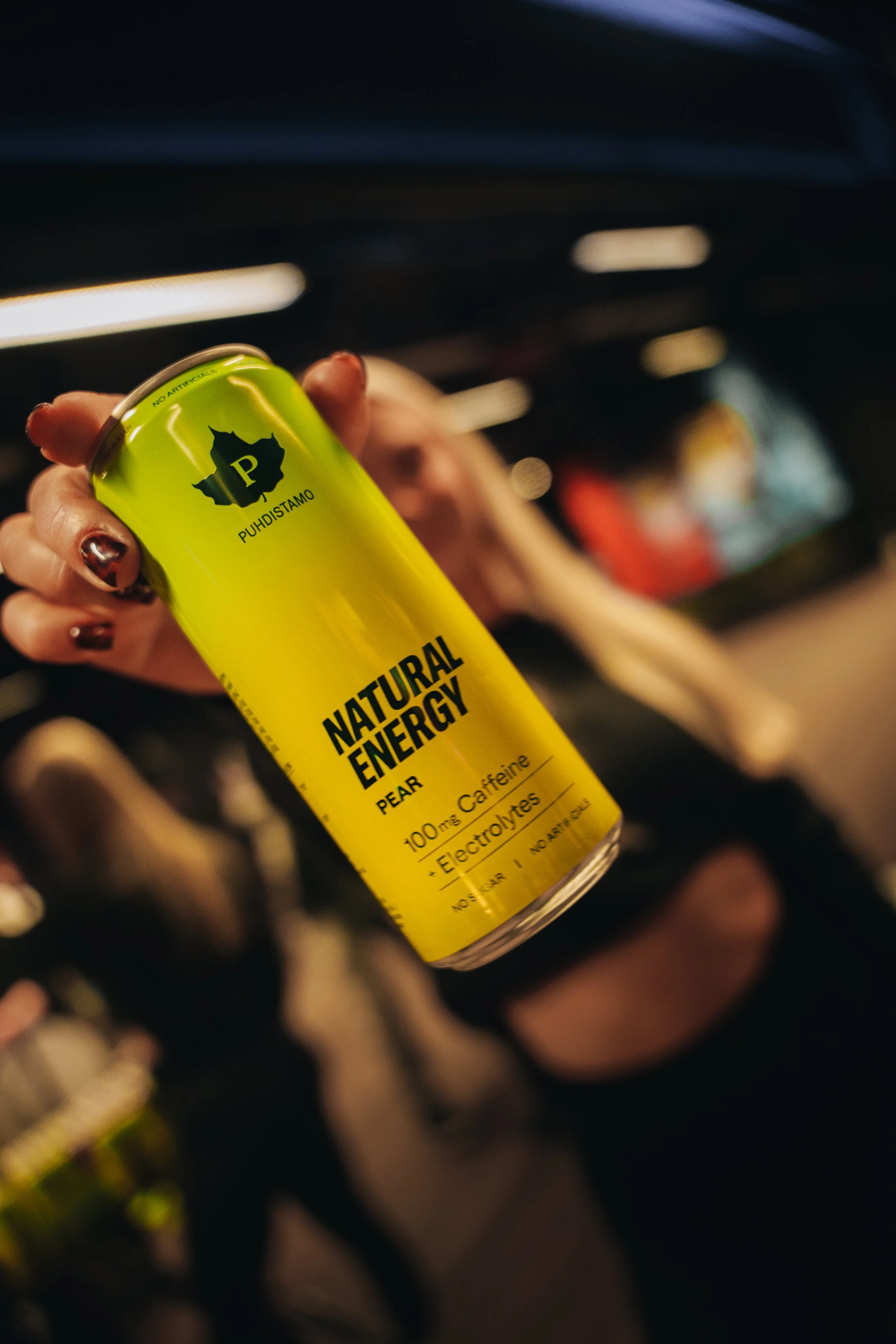 Kuva käsivarrest, joka pitää keltaista energiajuomasarjaa, jossa lukee 'Natural Energy' ja 'Pear', ja taustalla on pimeä sisätila.
