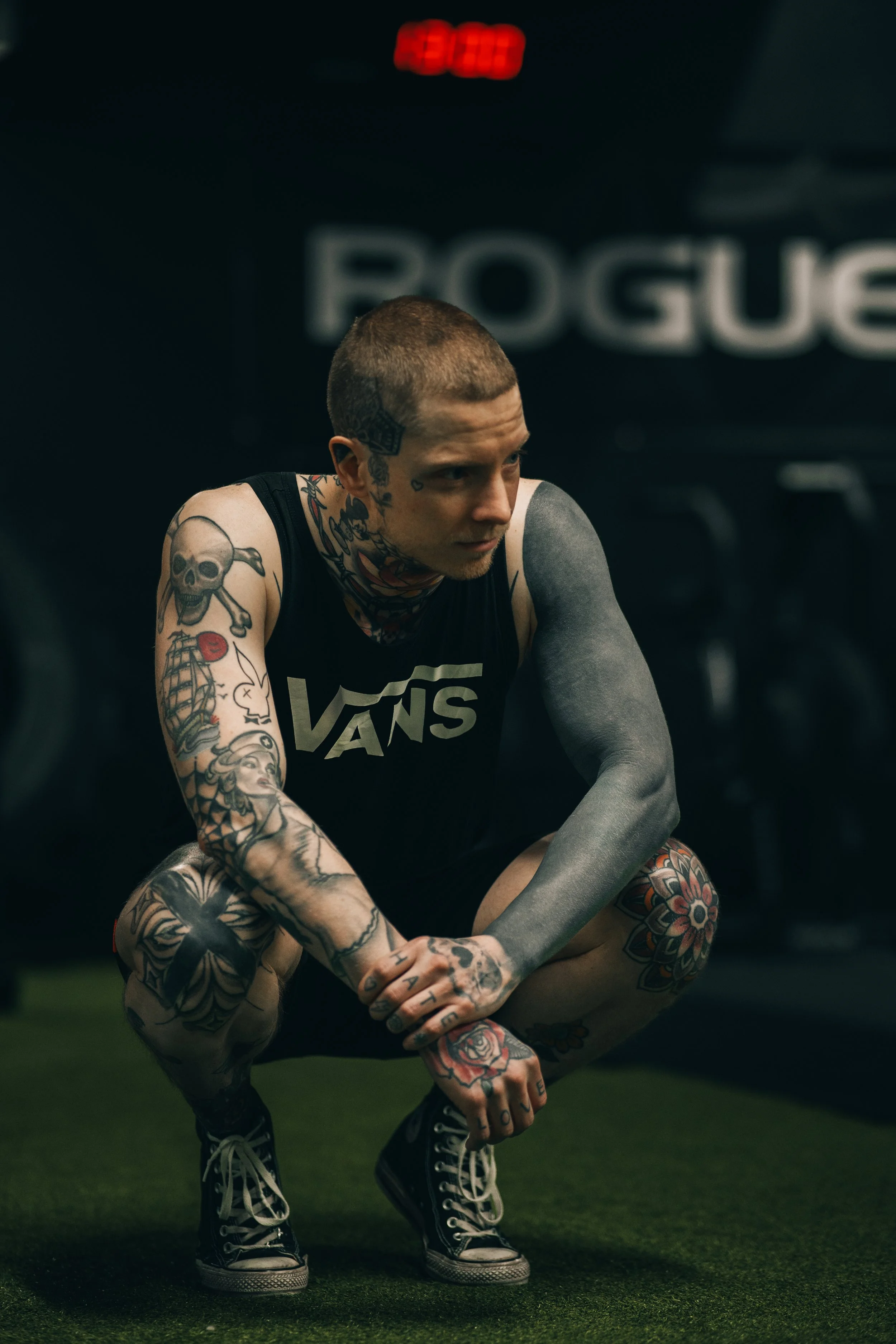 Nuori mies, jolla on paljon tatuointeja, istuu lattialla urheiluhallissa, taustalla näkyy teksti "ROGUE" ja digitaalinen kello.