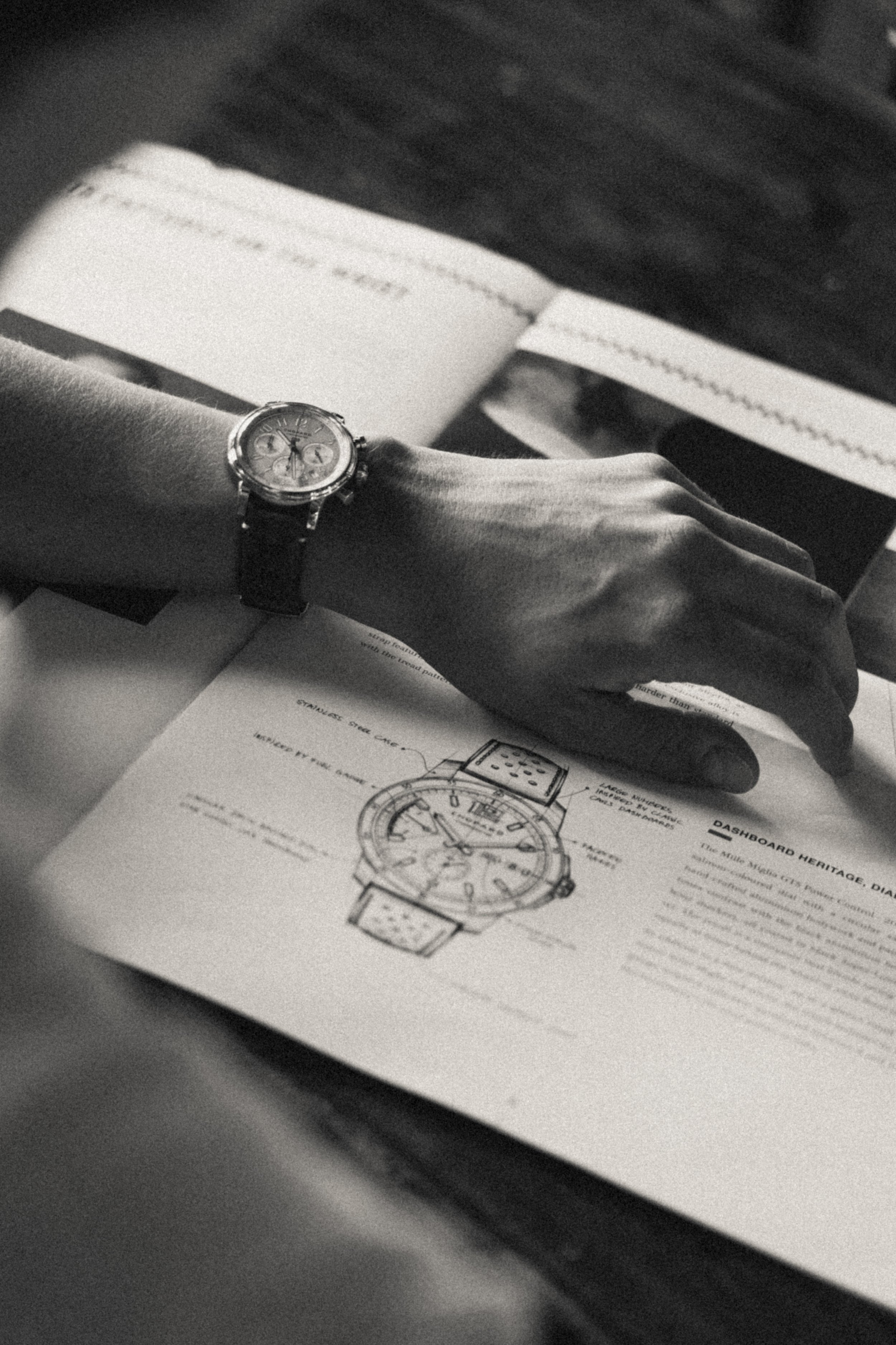 Une main portant une montre-bracelet posée sur un manuel ou un catalogue de montre, avec un dessin technique d'une montre. En arrière-plan, il y a des papiers ou des notes.