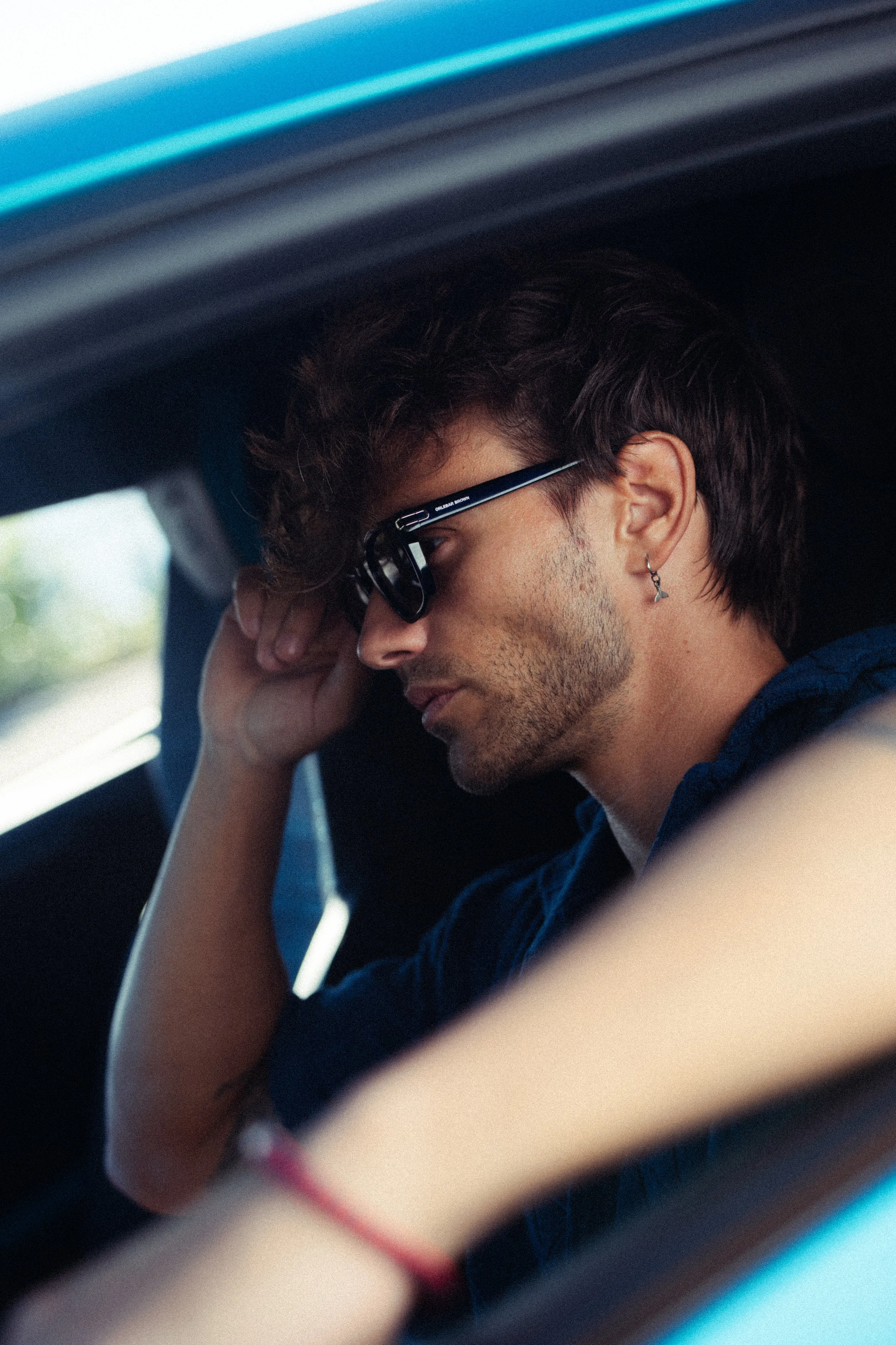 Un jeune homme avec des cheveux bouclés, portant des lunettes de soleil et une boucle d'oreille en forme de croix, repose sa tête sur sa main à l'intérieur d'une voiture