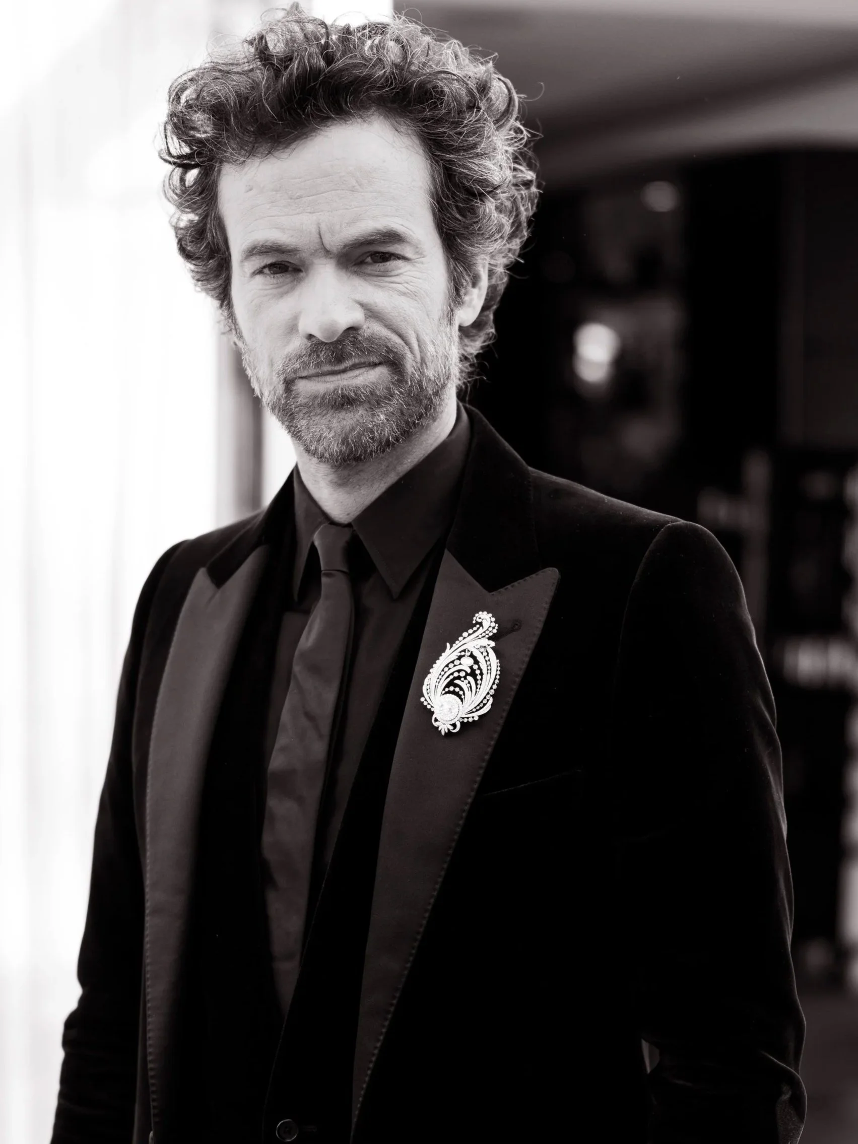 Un homme avec des cheveux bouclés et une barbe, portant un costume noir avec une broche argentée, dans un cadre intérieur.