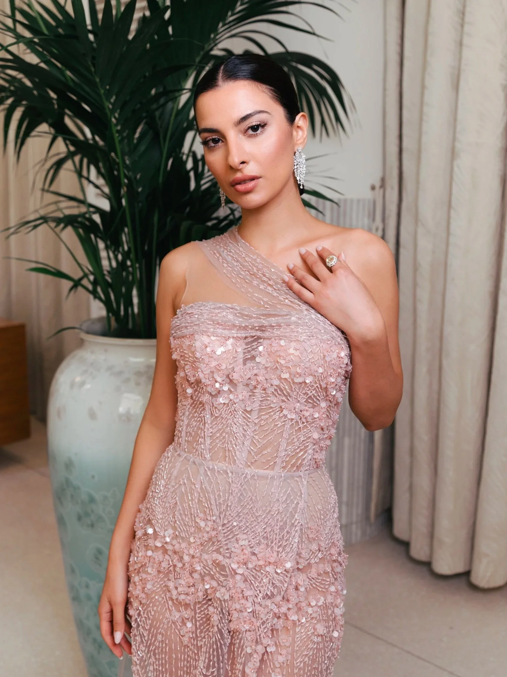 Jeune femme avec une robe rose pâle en dentelle et paillettes, portant des bijoux brillants, dans un environnement intérieur avec des plantes et des panneaux en bois.