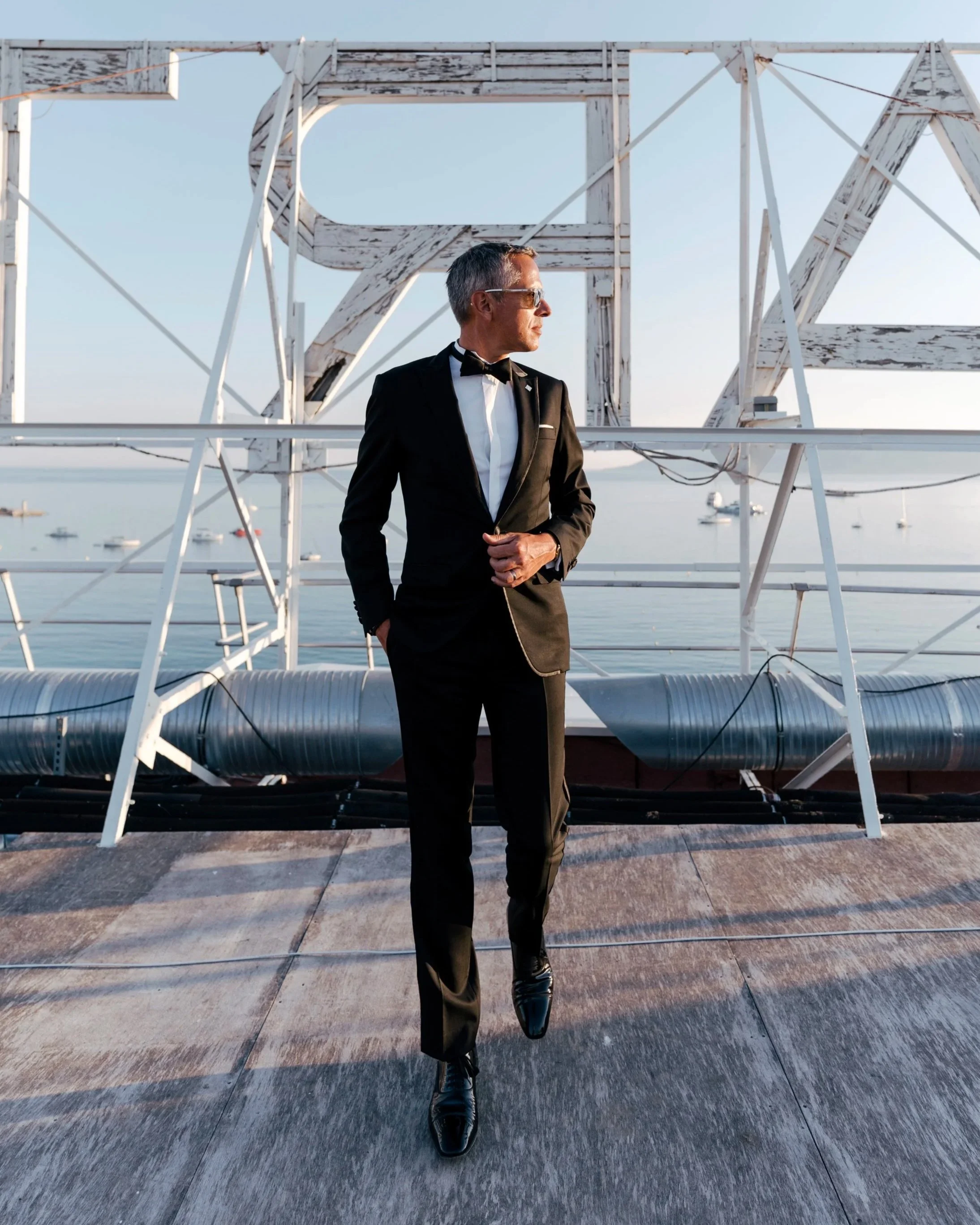 Homme élégant en smoking noir, portant des lunettes de soleil, debout sur un toit au bord de l'eau, avec une structure métallique en arrière-plan.