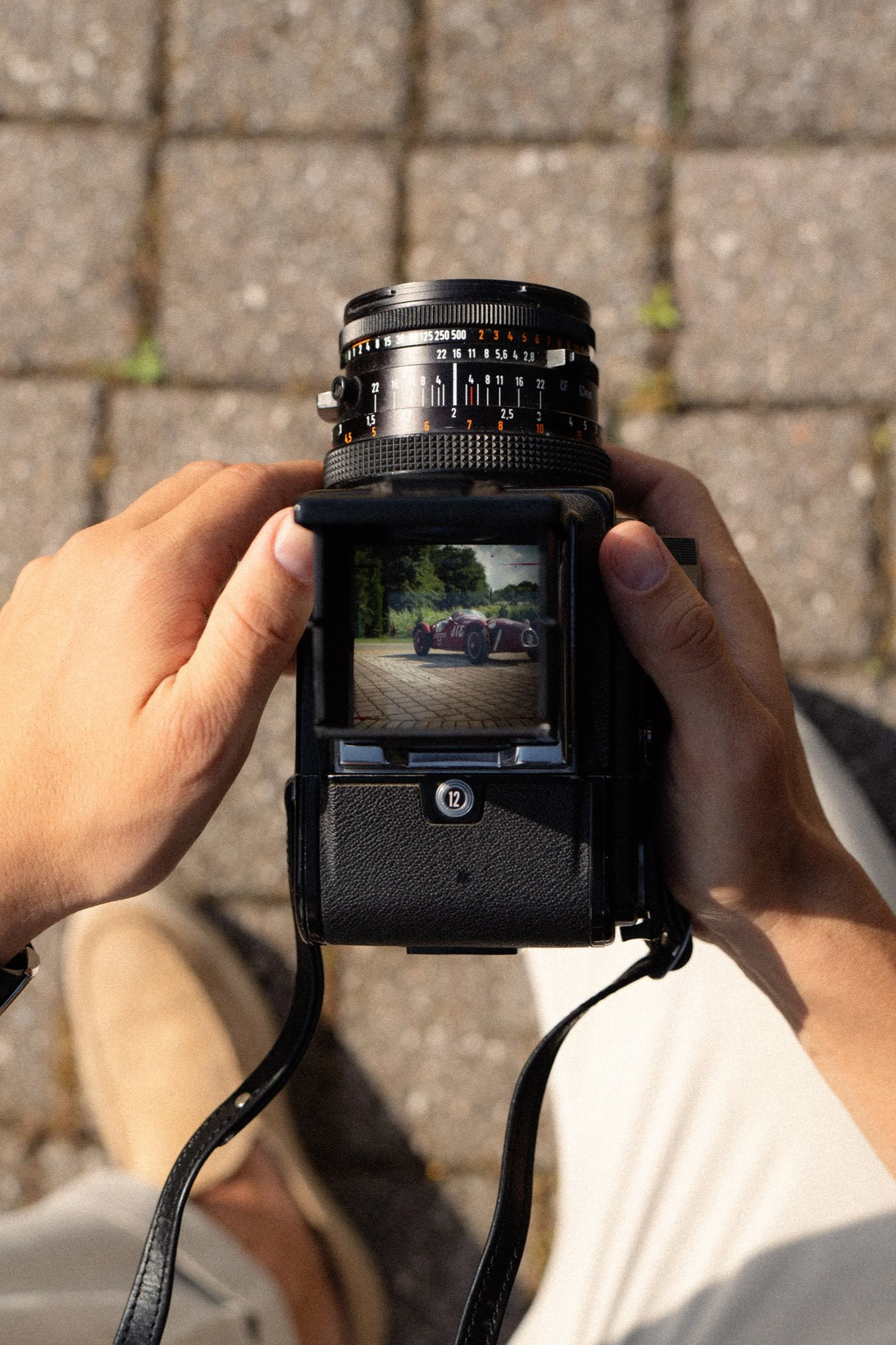 Une personne tient un appareil photo reflex en cadrant une voiture de course rouge, visible à l'écran de l'appareil, sur un sol pavé extérieur.