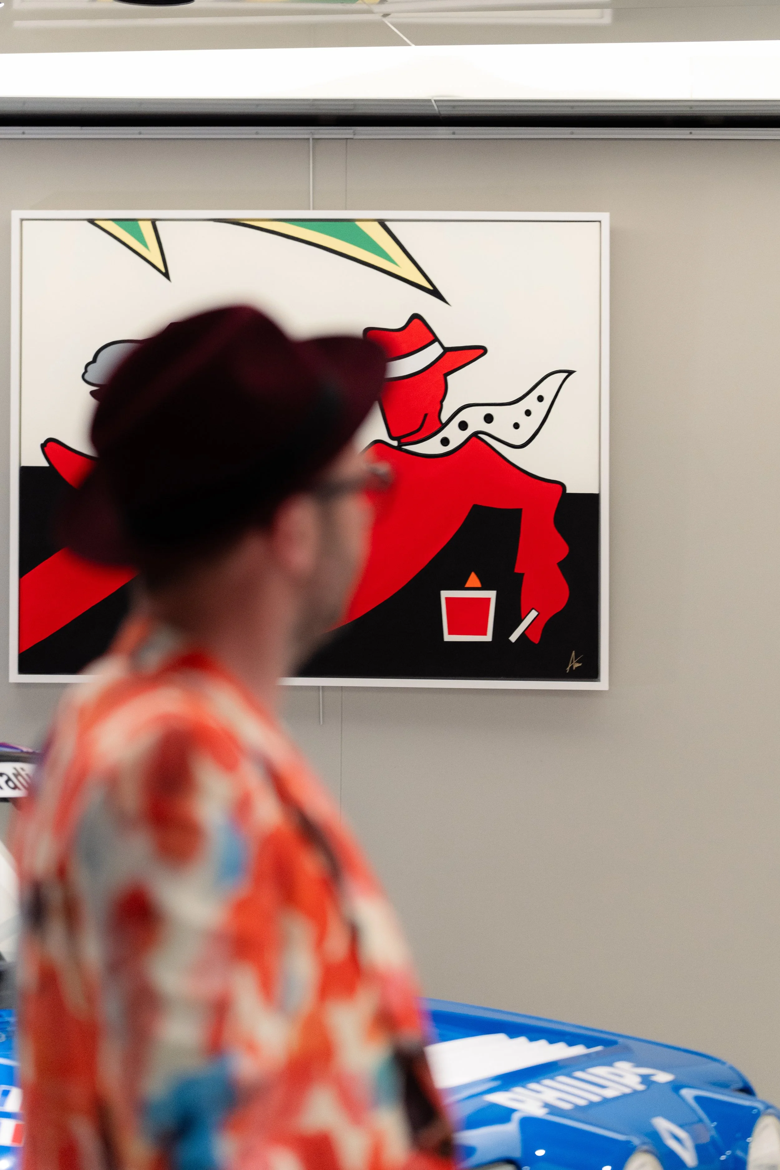 Une personne portant un chapeau rouge, une chemise colorée, et des lunettes est floue en premier plan, tandis qu'une œuvre d'art abstraite représentant une figure féminine stylisée en rouge, noir, blanc et beige est visible en arrière-plan.