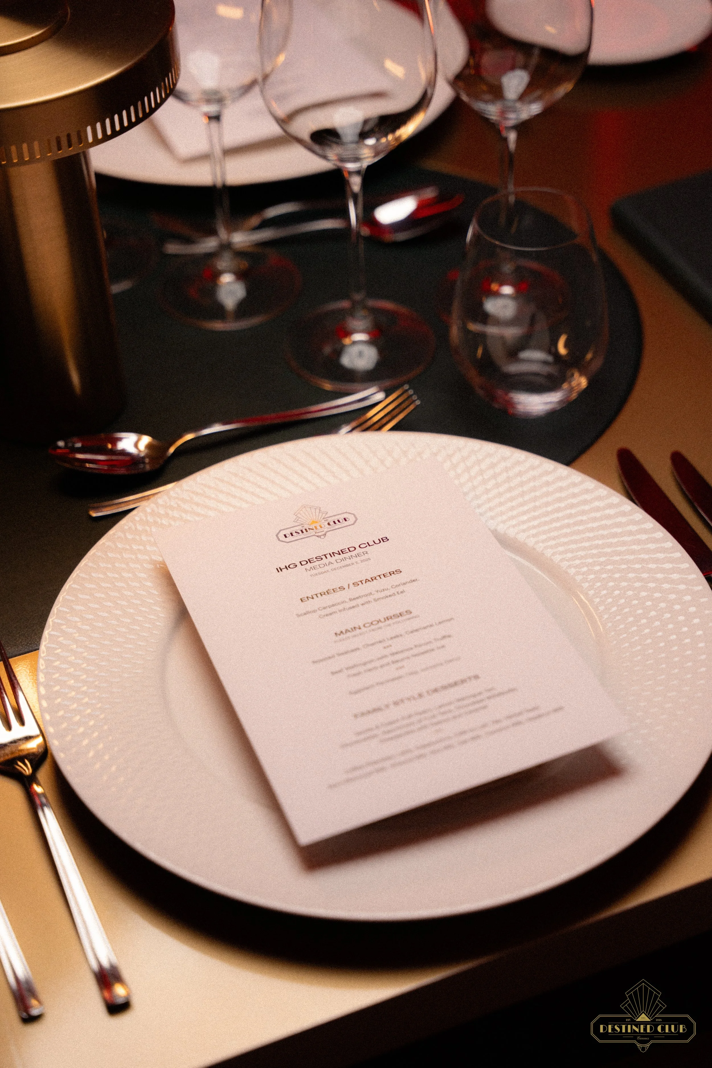 Une table élégante avec une assiette blanche, un menu, des couverts en métal, plusieurs verres à vin, et une lampe, dans un cadre de restaurant sophistiqué.