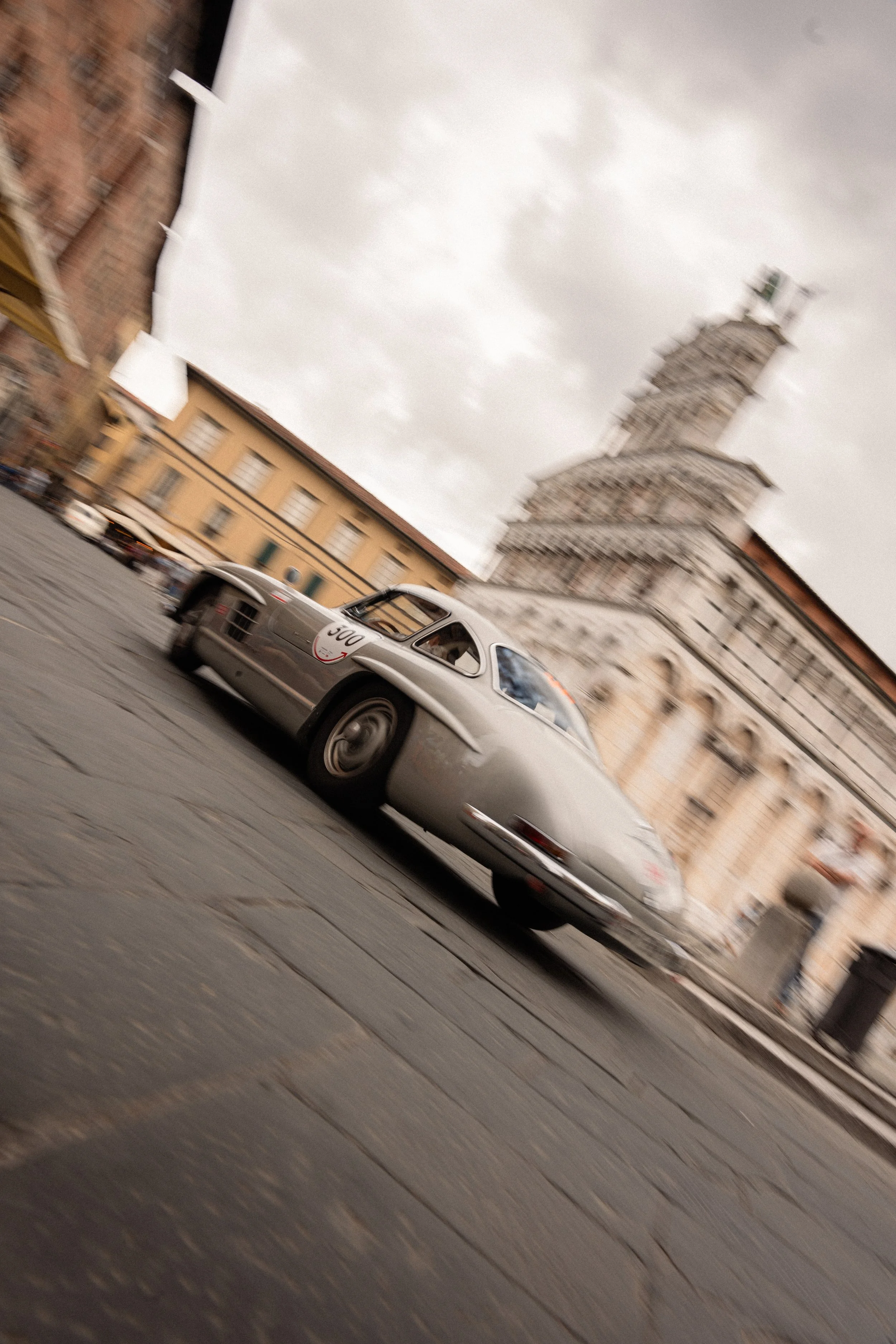 Voiture de course vintage argentée roulant sur une place pavée avec une tour penchée en arrière-plan, sous un ciel nuageux.