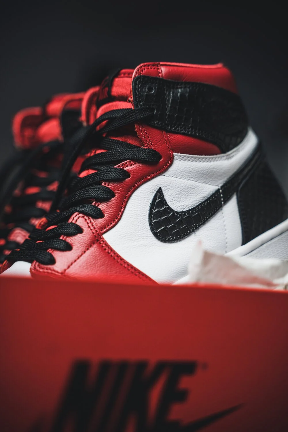 Une paire de baskets Nike Jordan 1 Retro High OG en noir, rouge, blanc, avec le logo noir en forme de Swoosh, dans une boîte orange Nike.