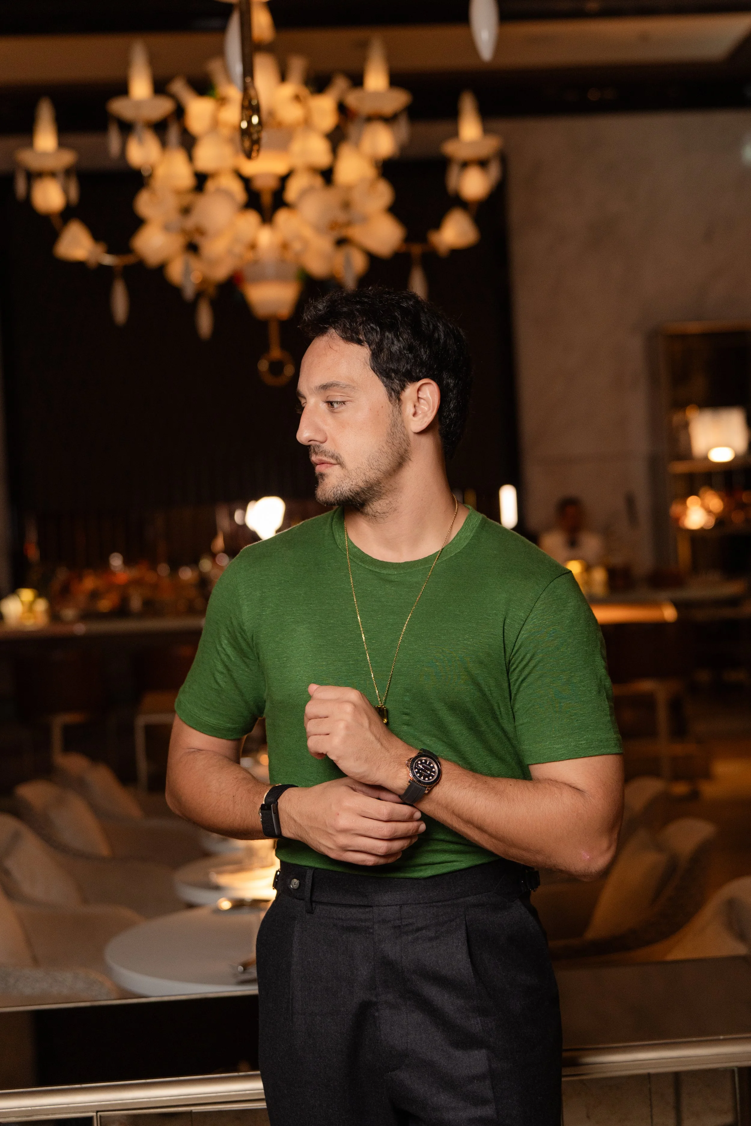 Un homme portant un t-shirt vert, une montre et un collier se trouve dans un restaurant élégant.